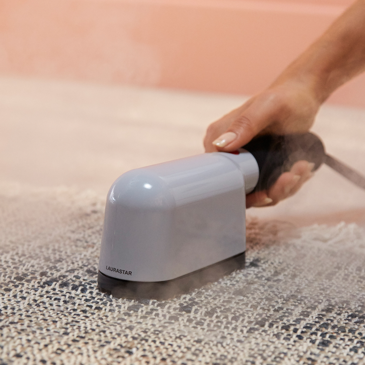 A hand holds a gray steam cleaner over a textured rug. Steam rises. A pink wall is in the background.