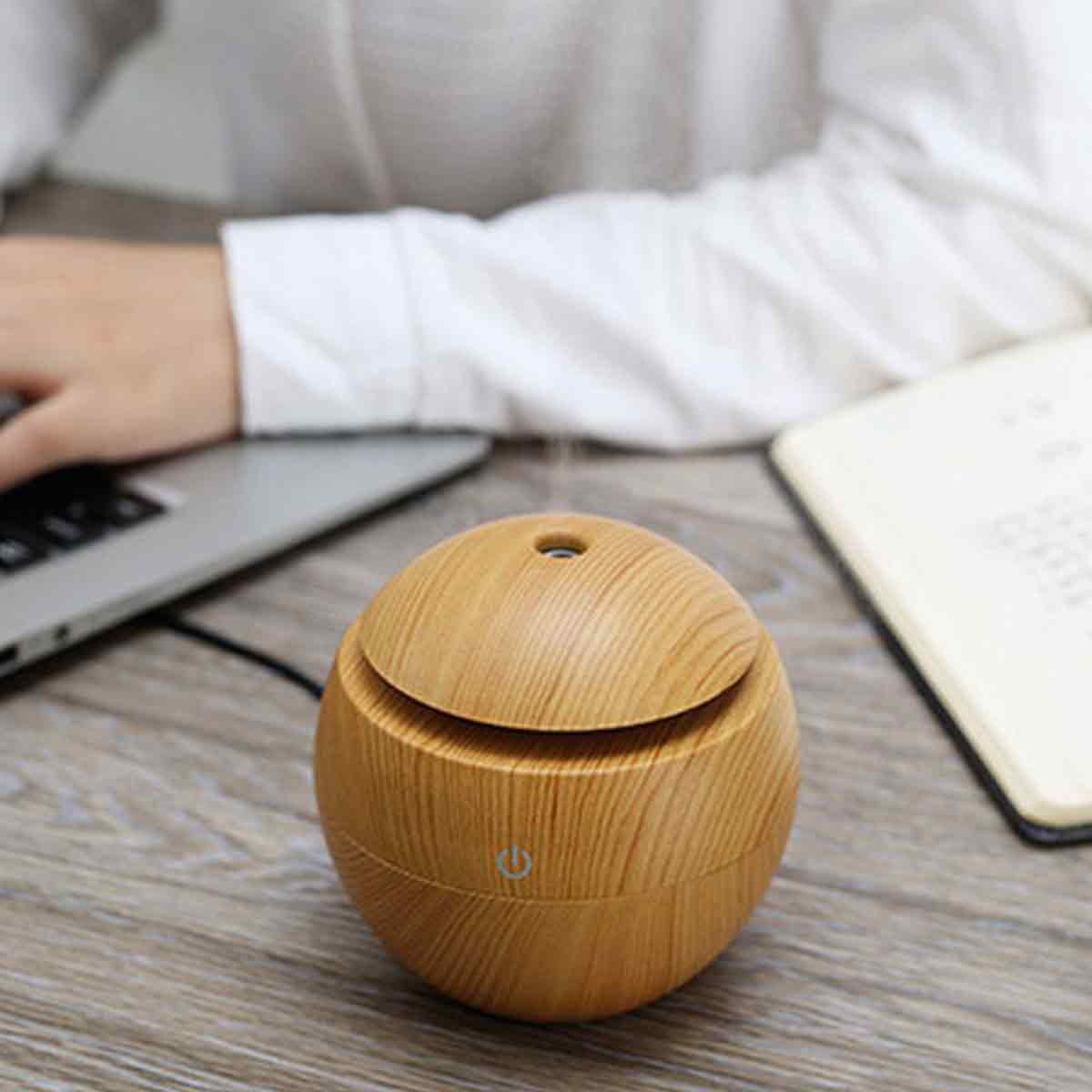 Ein hölzerner Diffusor für ätherische Öle auf einem Holzschreibtisch. Im Hintergrund arbeitet eine Person an einem Laptop.