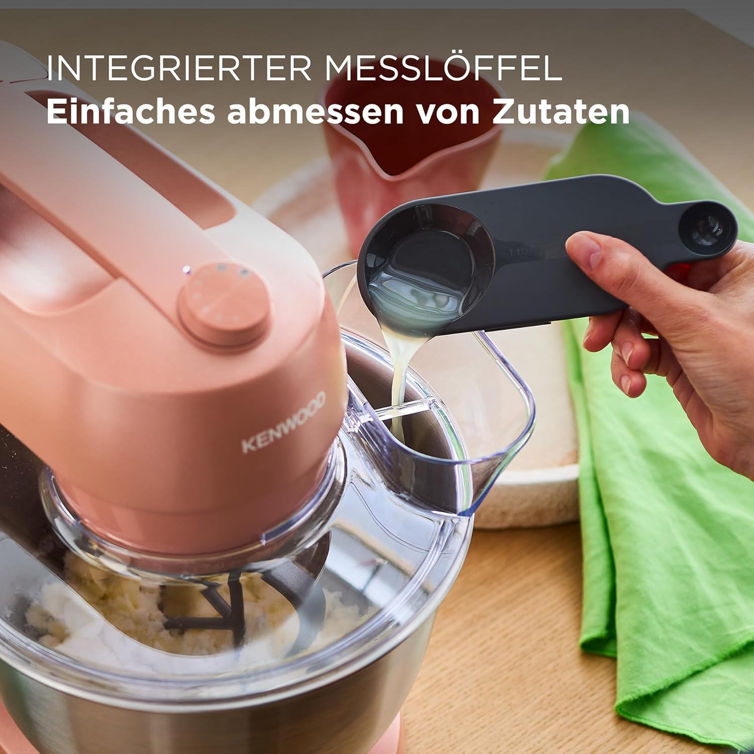 Eine Hand gießt Flüssigkeit aus einem Messlöffel in einen Mixer. Ein rosa Mixer und ein grünes Tuch sind sichtbar.