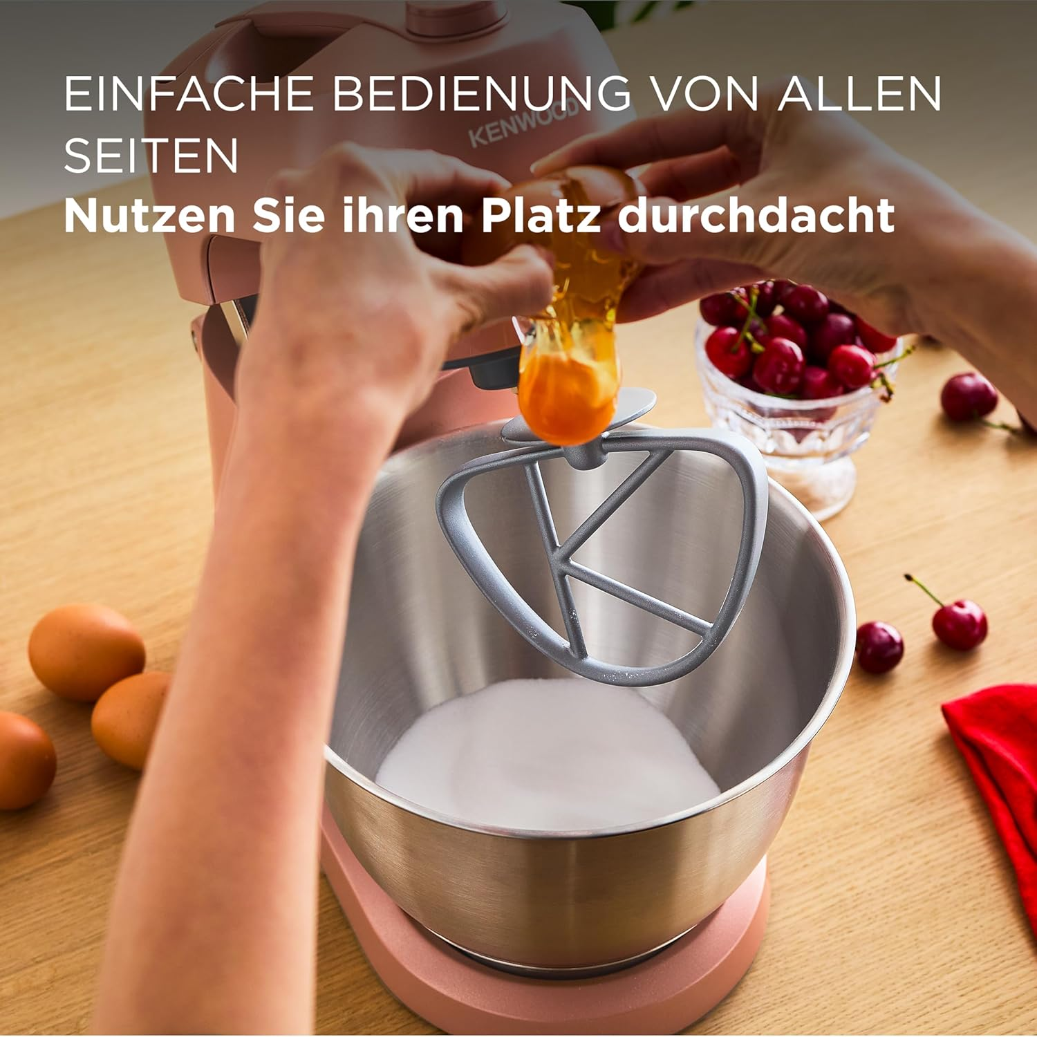 Eine Person, die ein Ei in eine Mixerschüssel mit Zucker schlägt. Kirschen und Eier liegen ebenfalls auf der Holzoberfläche.