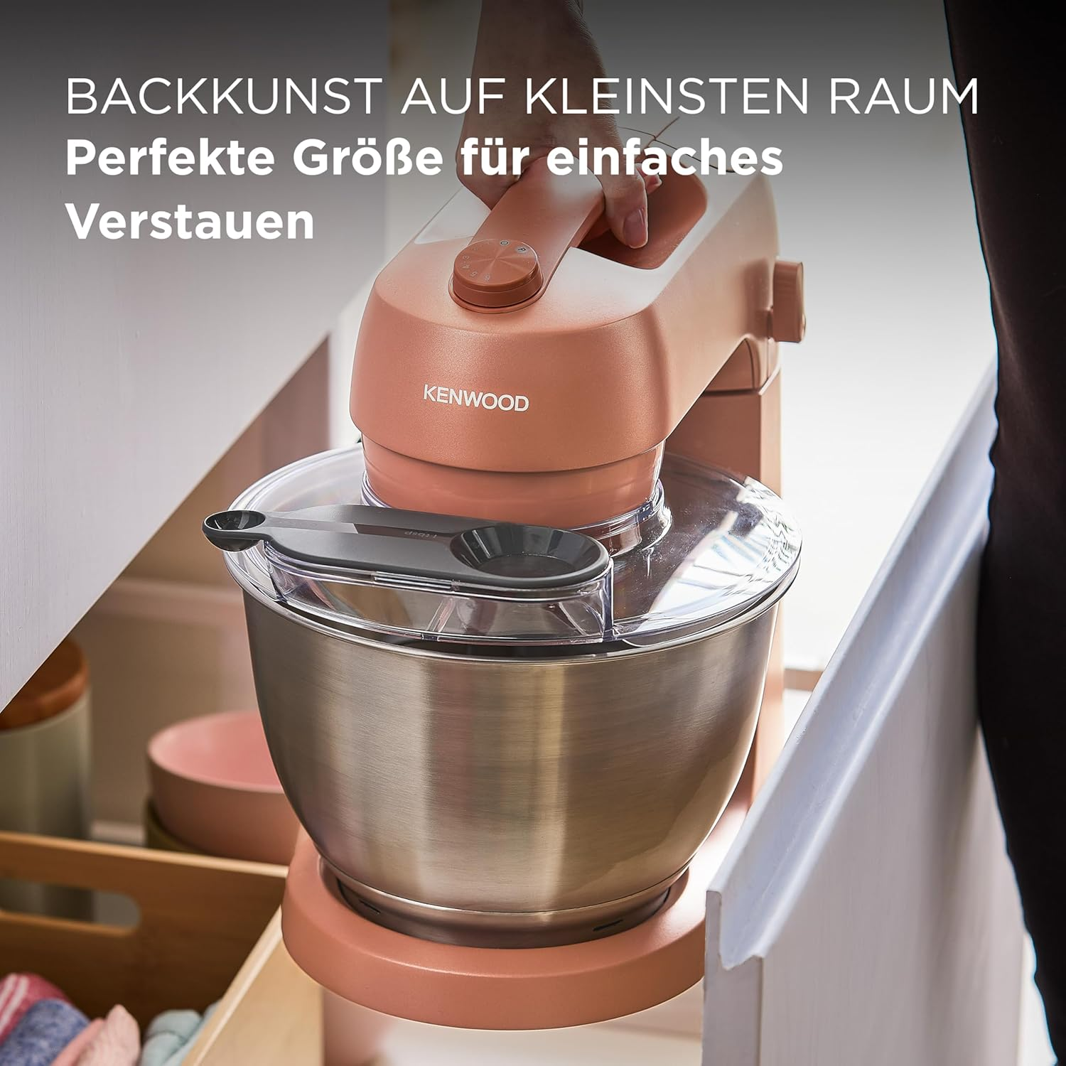 Ein pfirsichfarbener Kenwood-Küchenmixer mit einer Edelstahlschüssel steht auf einer Theke. Eine Hand befindet sich am Arm der Maschine.