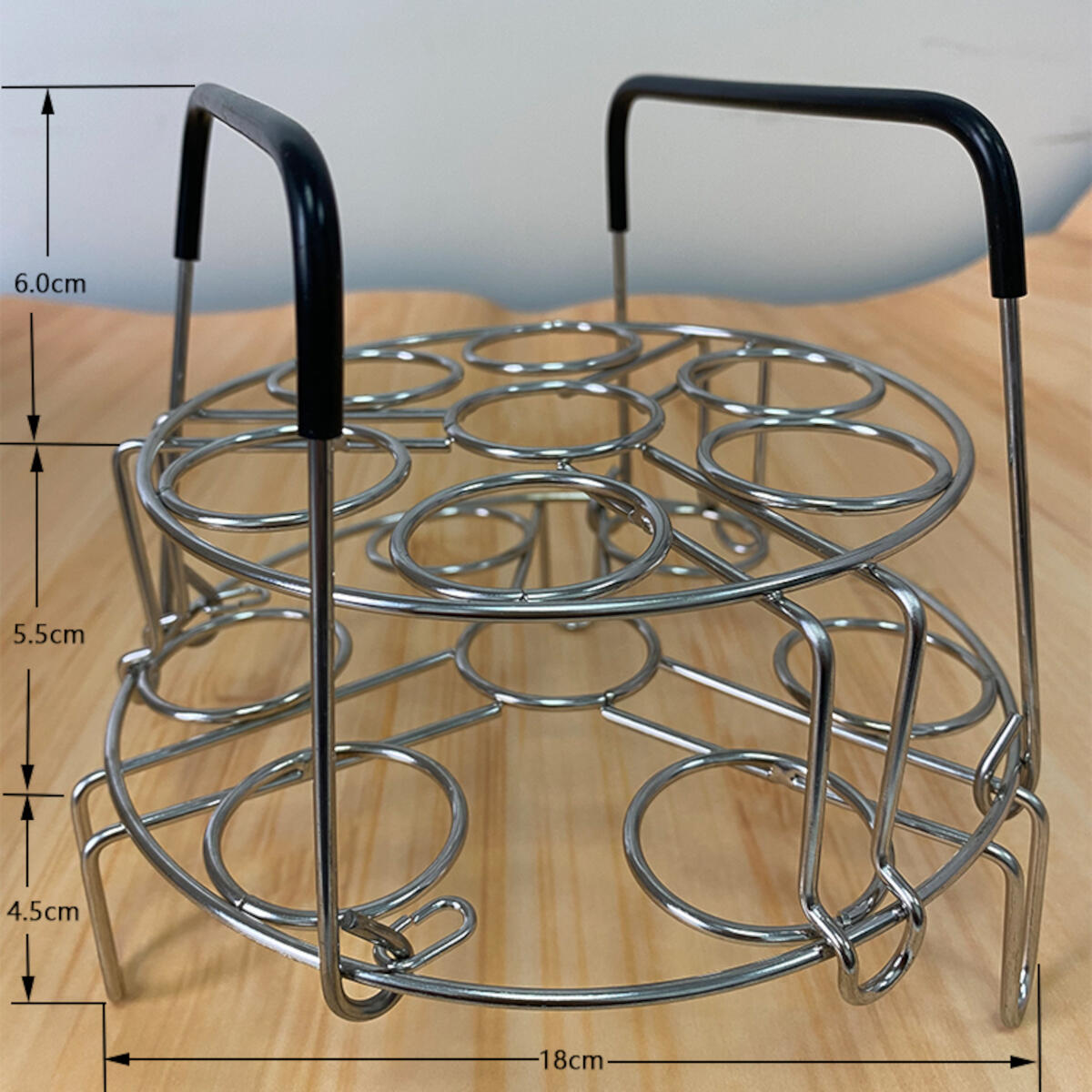 A metal egg rack with two levels and handles. It is placed on a wooden surface. Dimensions are indicated by arrows.