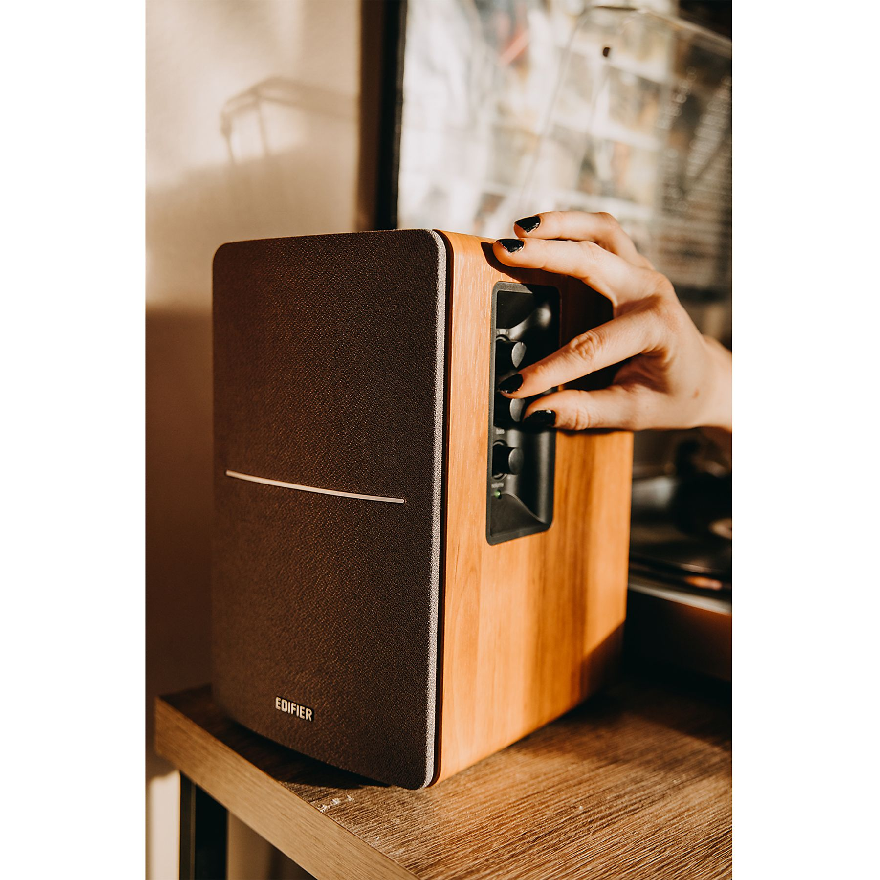 Ręka z czarnymi paznokciami reguluje elementy głośnika. Głośnik jest brązowy i w kolorze drewna, na drewnianej powierzchni.