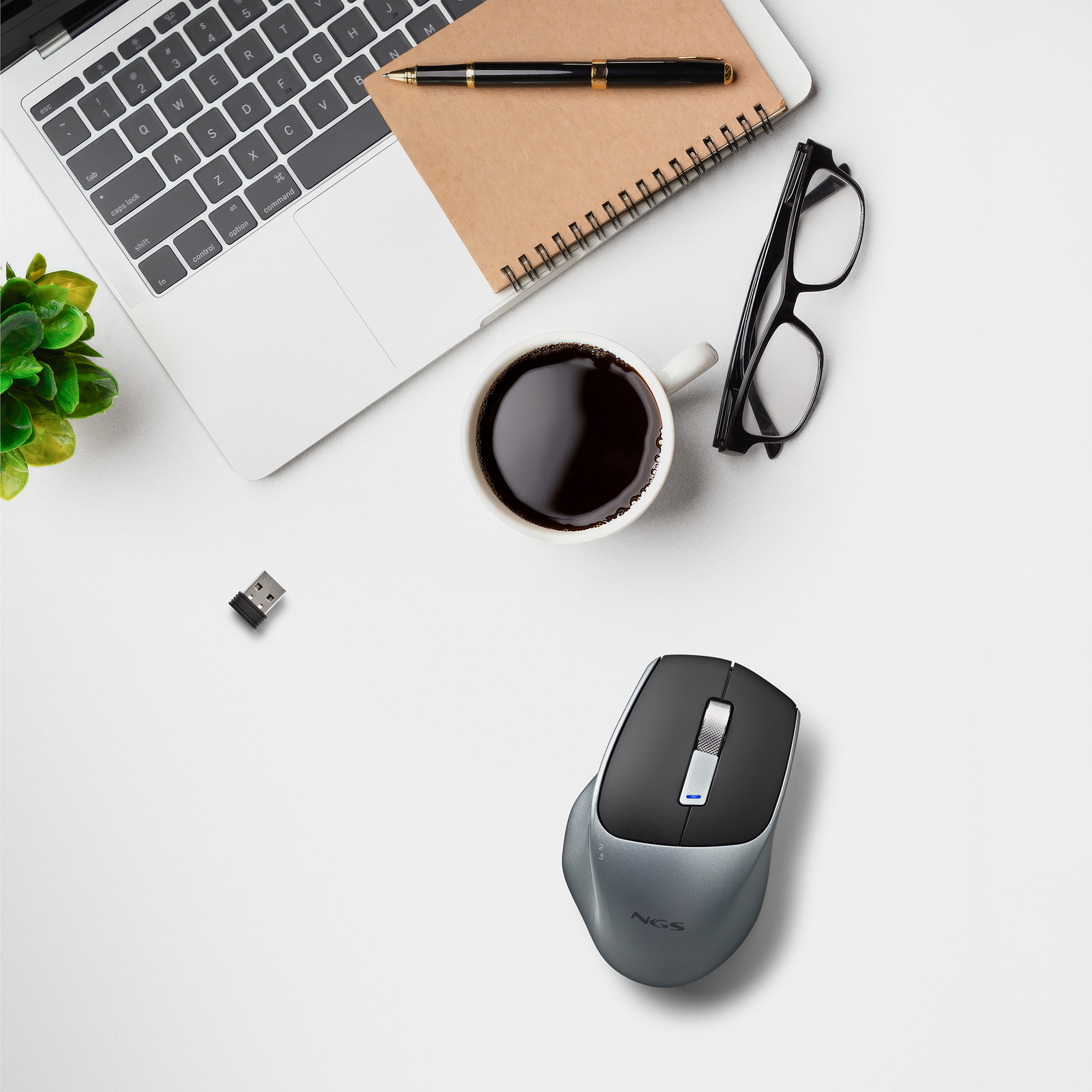 Una scrivania con laptop, caffè, occhiali, penna, blocco note, mouse e una piccola pianta, tutto su una superficie bianca.