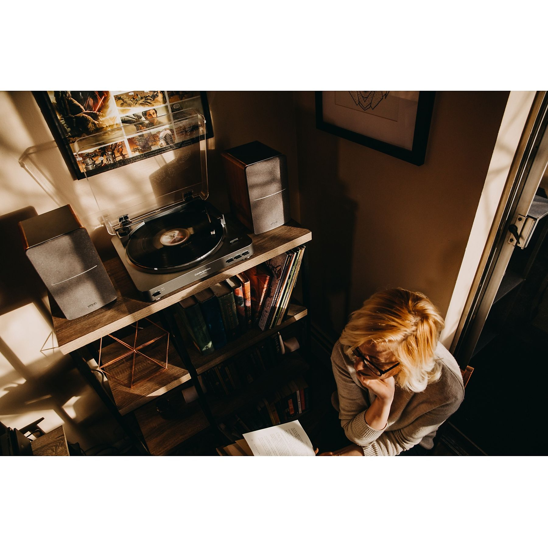Kobieta z blond włosami czyta, blisko gramofonu i półki na książki. Delikatne światło słoneczne wnika do środka.