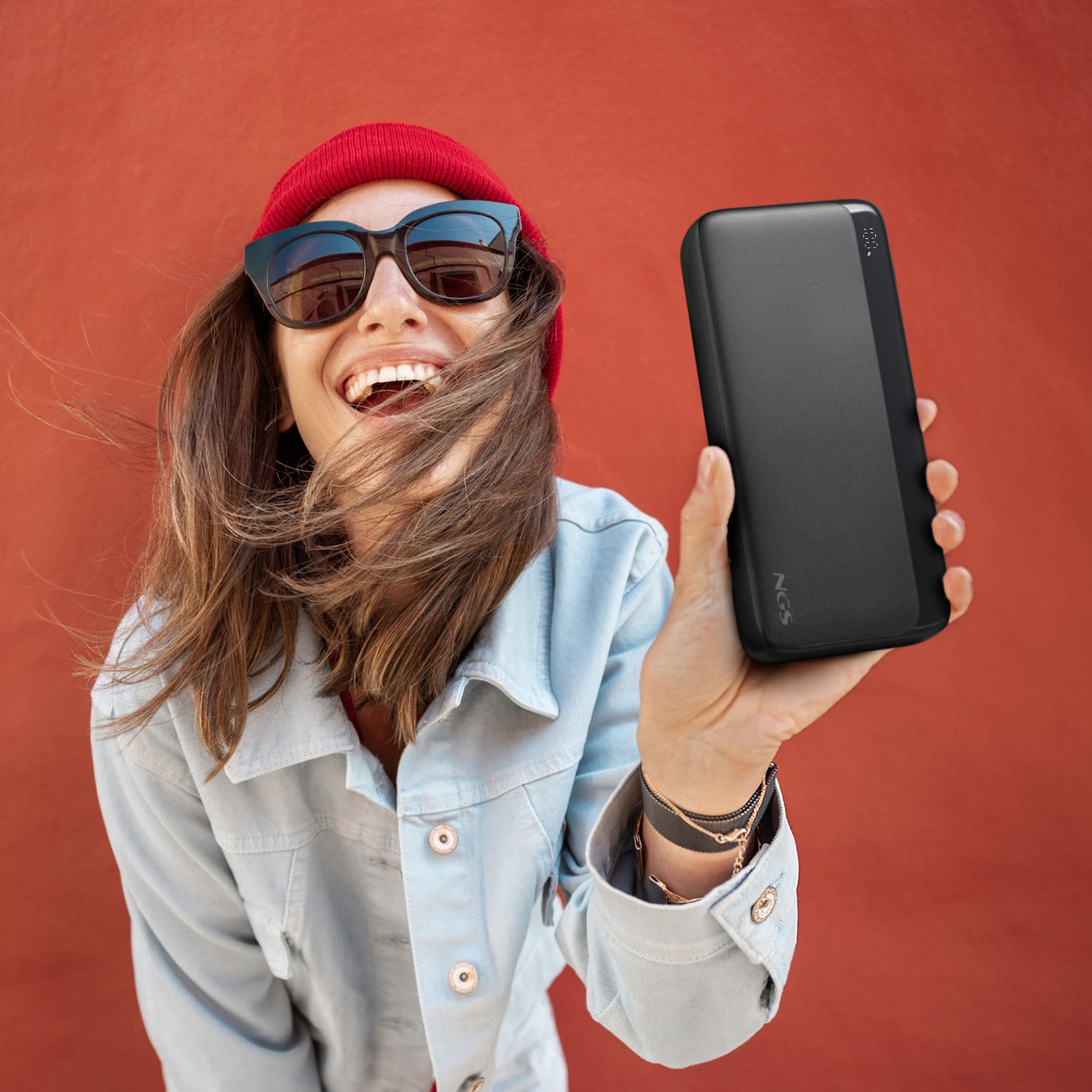 Una mujer sonriendo, sosteniendo un banco de energía negro. Lleva gafas de sol, gorro rojo, chaqueta vaquera y pulsera sobre fondo rojo.