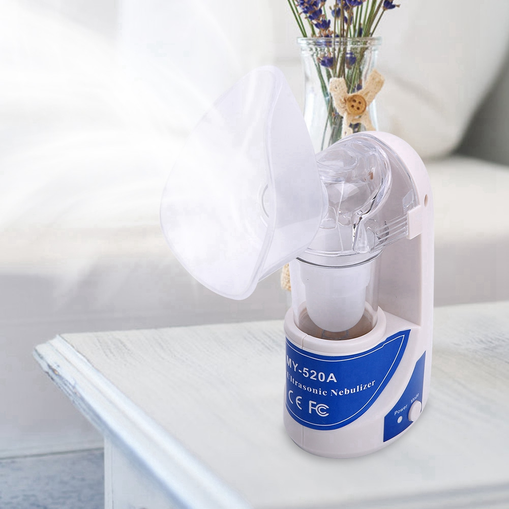 Ein weiß-blauer Ultraschallvernebler mit klarer Maske, eine Vase mit lila Blumen im Hintergrund, auf einem weißen Tisch.