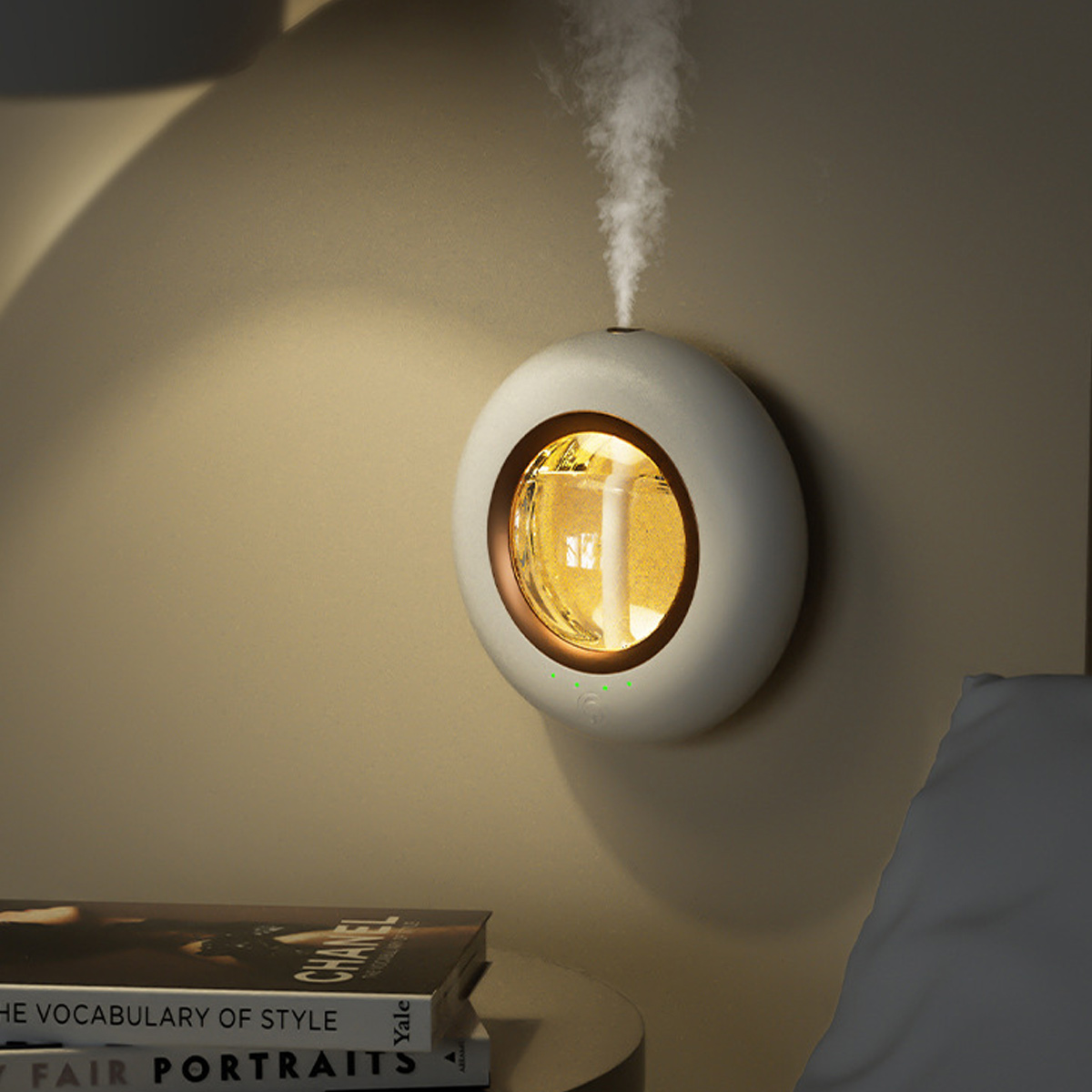 Ein weiß-goldener Diffusor für ätherische Öle an einer Wand, der Dampf abgibt. Bücher im Vordergrund, ein Kissen im Hintergrund.