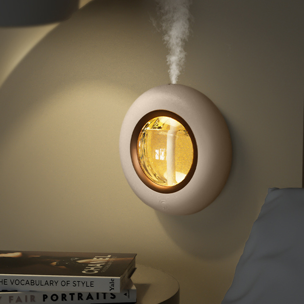 Ein beiger Diffusor, der Dampf abgibt. Er hat eine runde Form mit goldenem Innenlicht. Ein Buch und ein Bett sind teilweise zu sehen.
