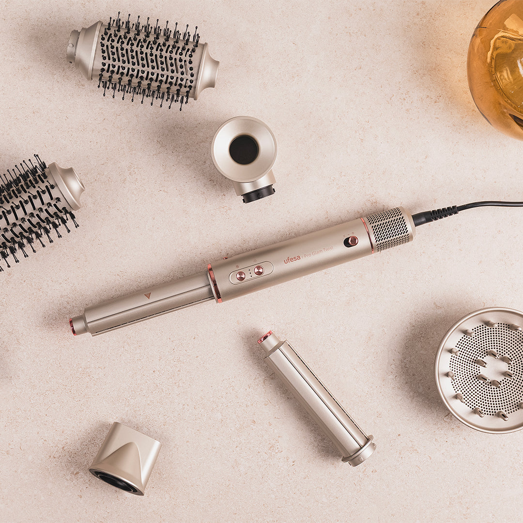 Strumenti per lo styling dei capelli disposti su una superficie chiara. Include un asciugacapelli, accessori per spazzole e un diffusore. Dominano i colori beige e metallico.