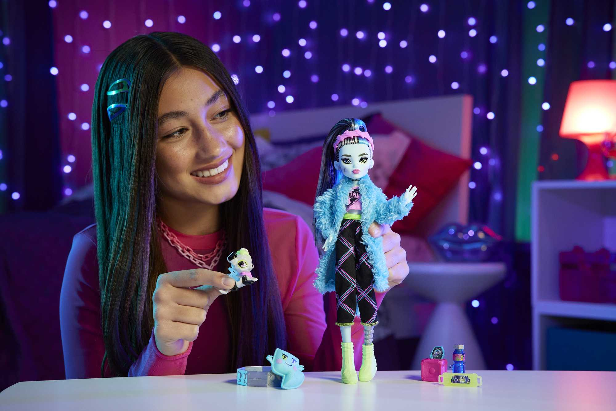 Una chica sonríe sosteniendo una muñeca y una pequeña figura. La muñeca tiene cabello azul y un abrigo azul. El fondo tiene luces púrpuras.
