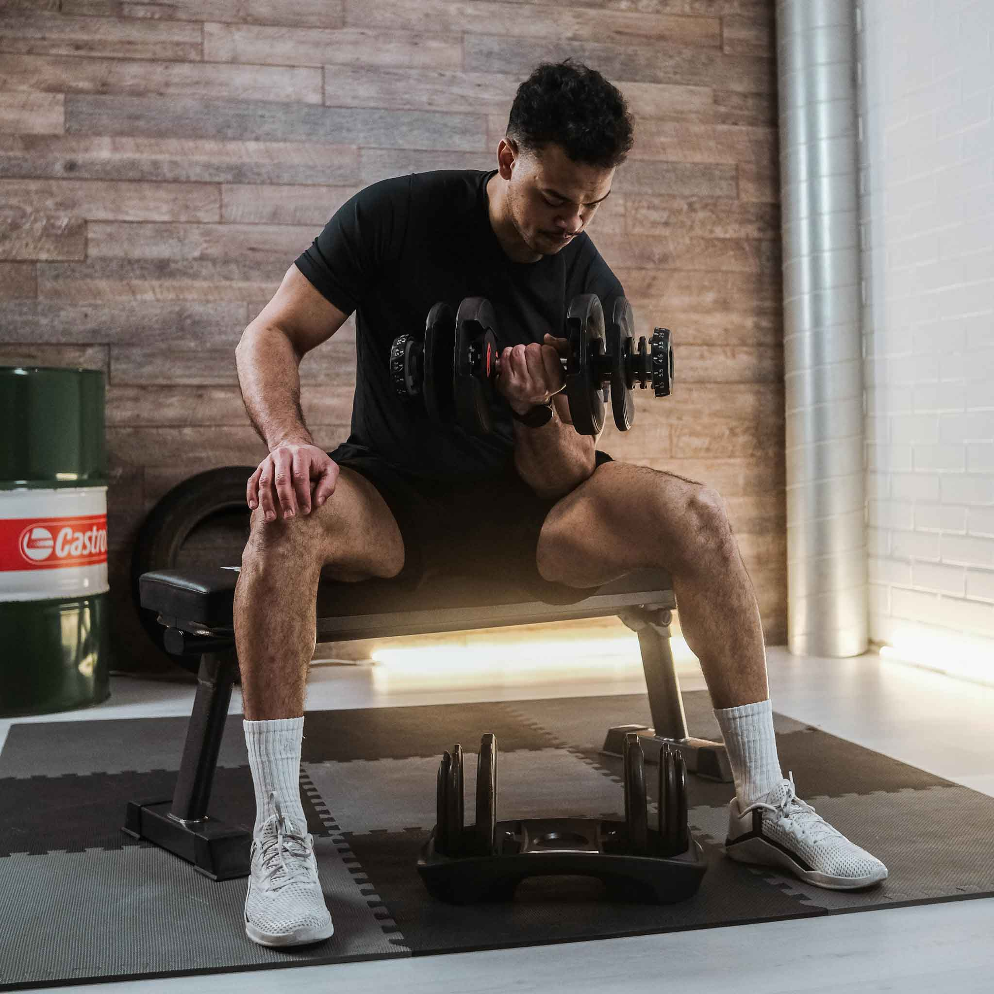 Mann in Schwarz, der eine Hantel hebt, auf einer Bank sitzt. Fitnessstudio mit Gewichten und Fliesen.