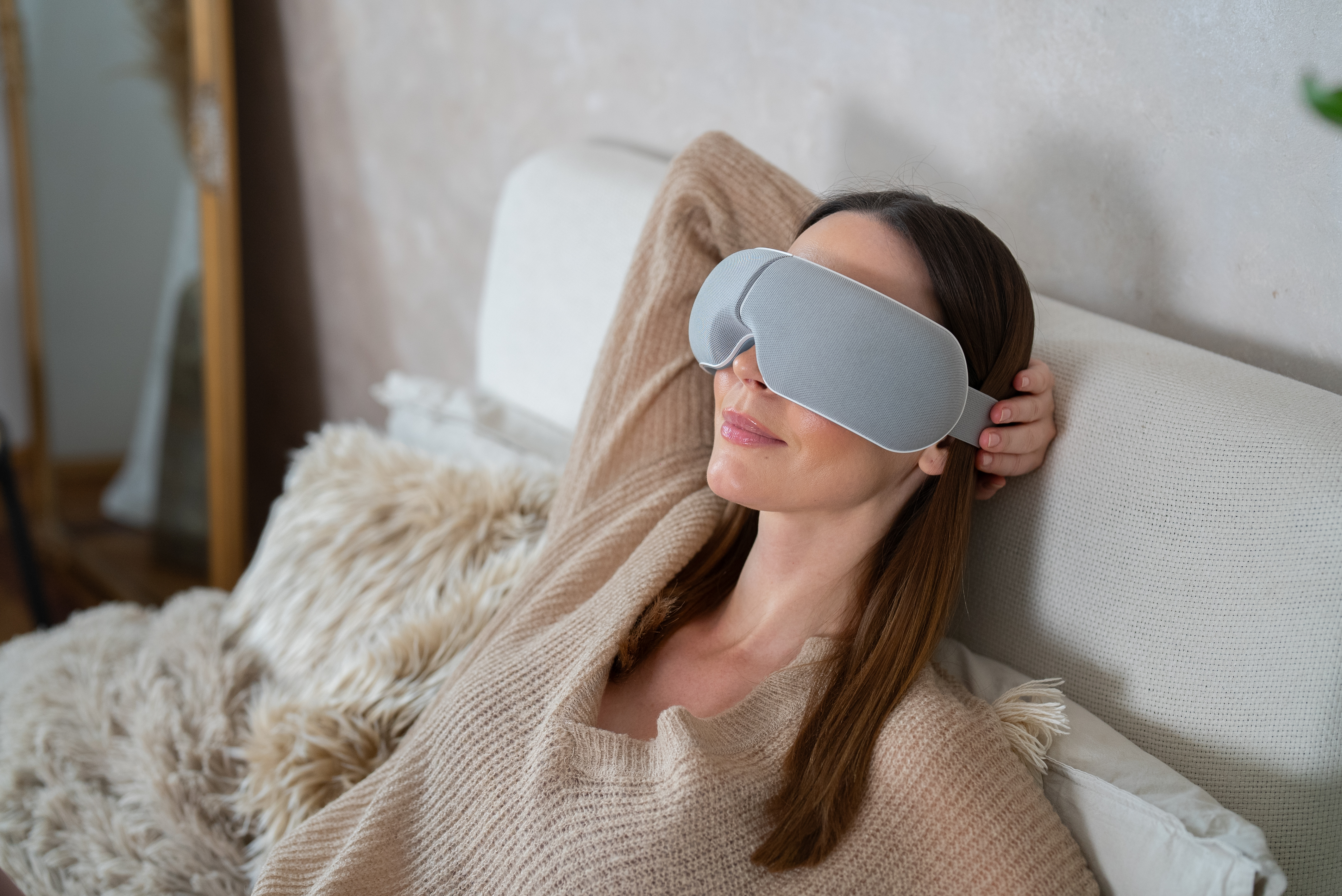 Mujer con antifaz durmiendo en cama. Suéter beige, almohada peluda y espejo son visibles.