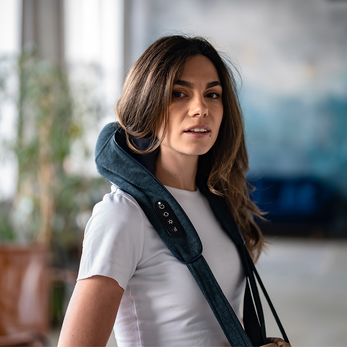 Una mujer con un masajeador de cuello mira a la cámara. Lleva una camisa blanca. El fondo está borroso.