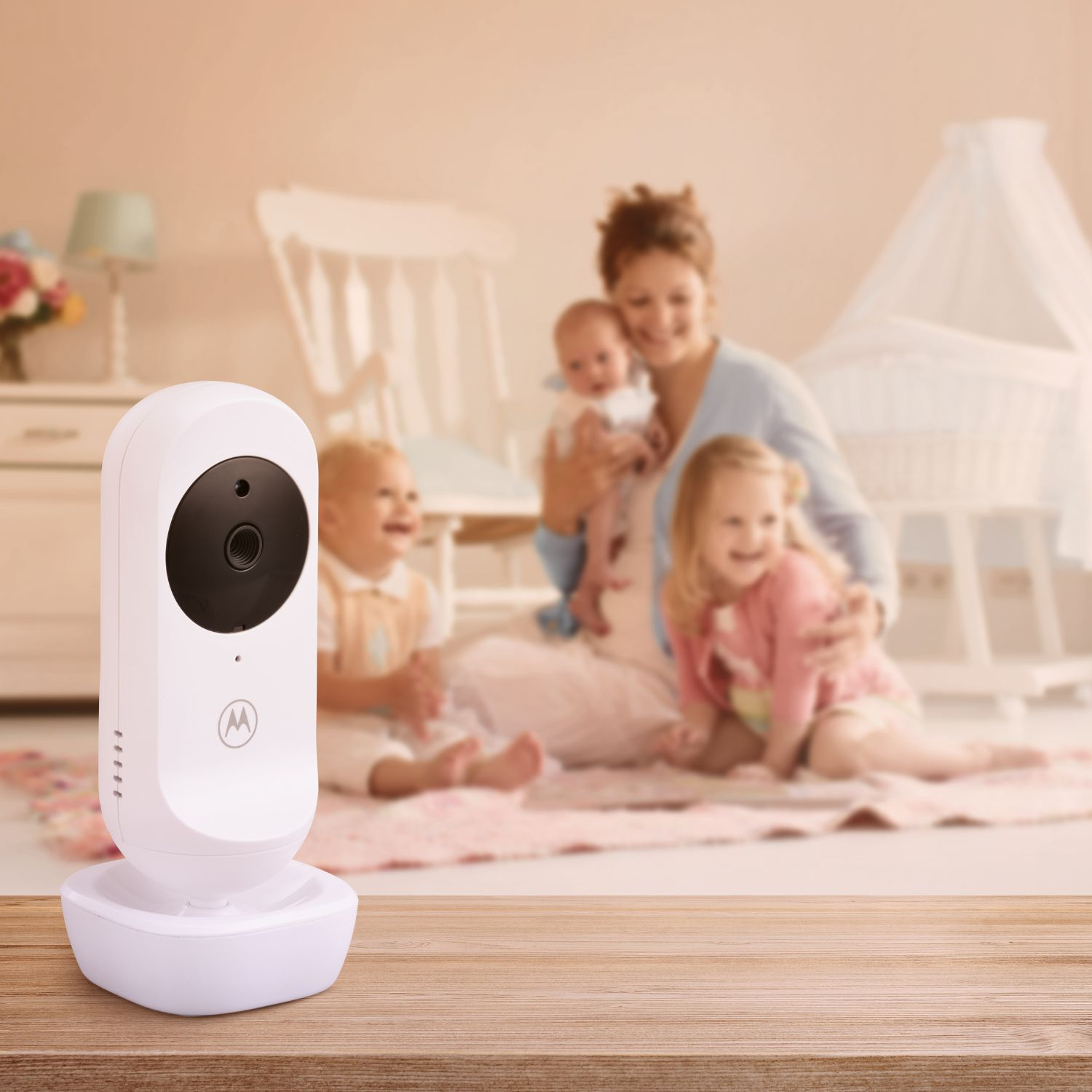 Ein weißer Babyphone steht auf einem Holztisch. Der unscharfe Hintergrund zeigt eine vierköpfige Familie in einem Kinderzimmer.