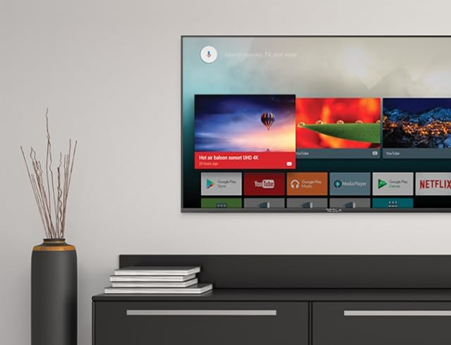 Una pantalla de TV muestra aplicaciones como YouTube y Netflix. Un jarrón con ramas secas se encuentra cerca del televisor y una pila de libros sobre un mueble.