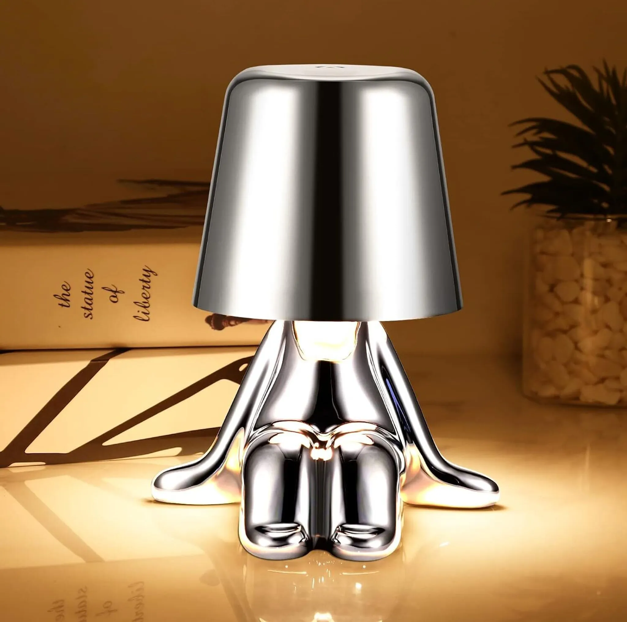 Eine verchromte Lampe in Form einer sitzenden Person mit Lampenschirm. Bücher mit Text und eine Pflanze im Hintergrund.
