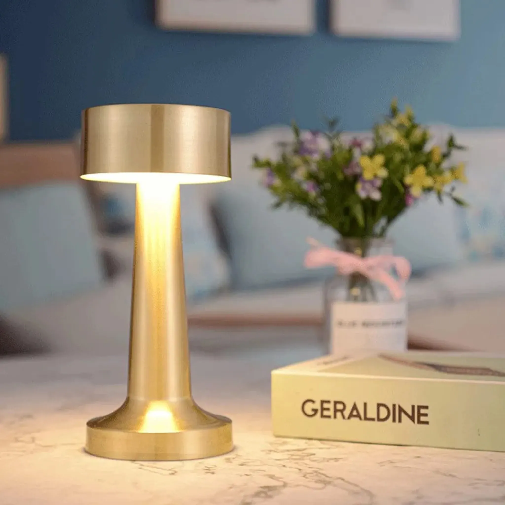 Auf einem Tisch steht eine goldene Lampe. Blumen und ein Buch sind im Hintergrund.