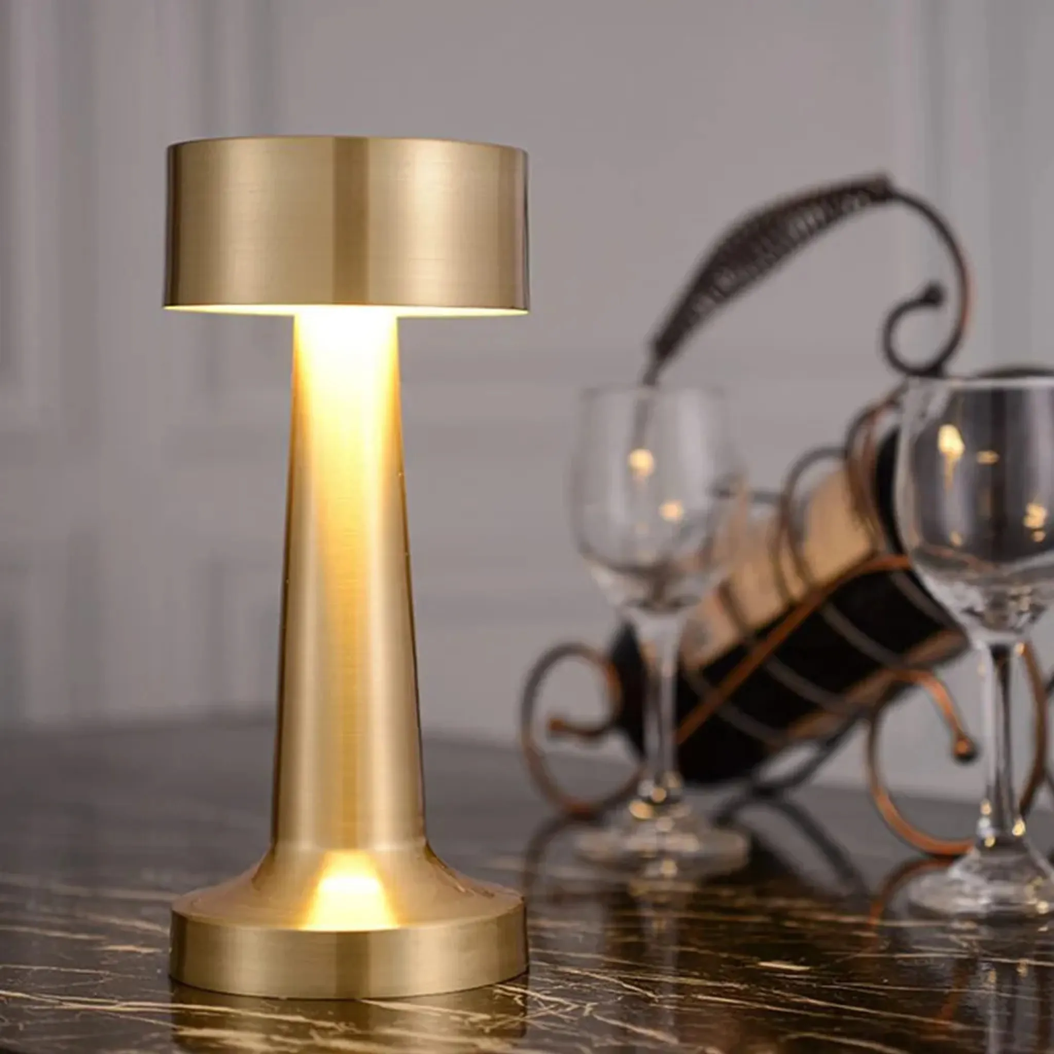 Eine goldene Lampe auf einem Tisch, mit Weingläsern und einem Gestell im Hintergrund. Die Lampe ist an.