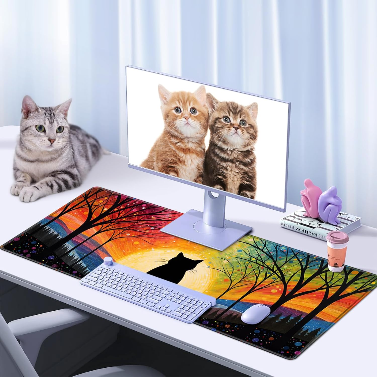 Schreibtisch mit Katze, Monitor mit Kätzchen und farbenfroher Mauspad mit Sonnenuntergang und Bäumen. Eine andere Katze sitzt links.