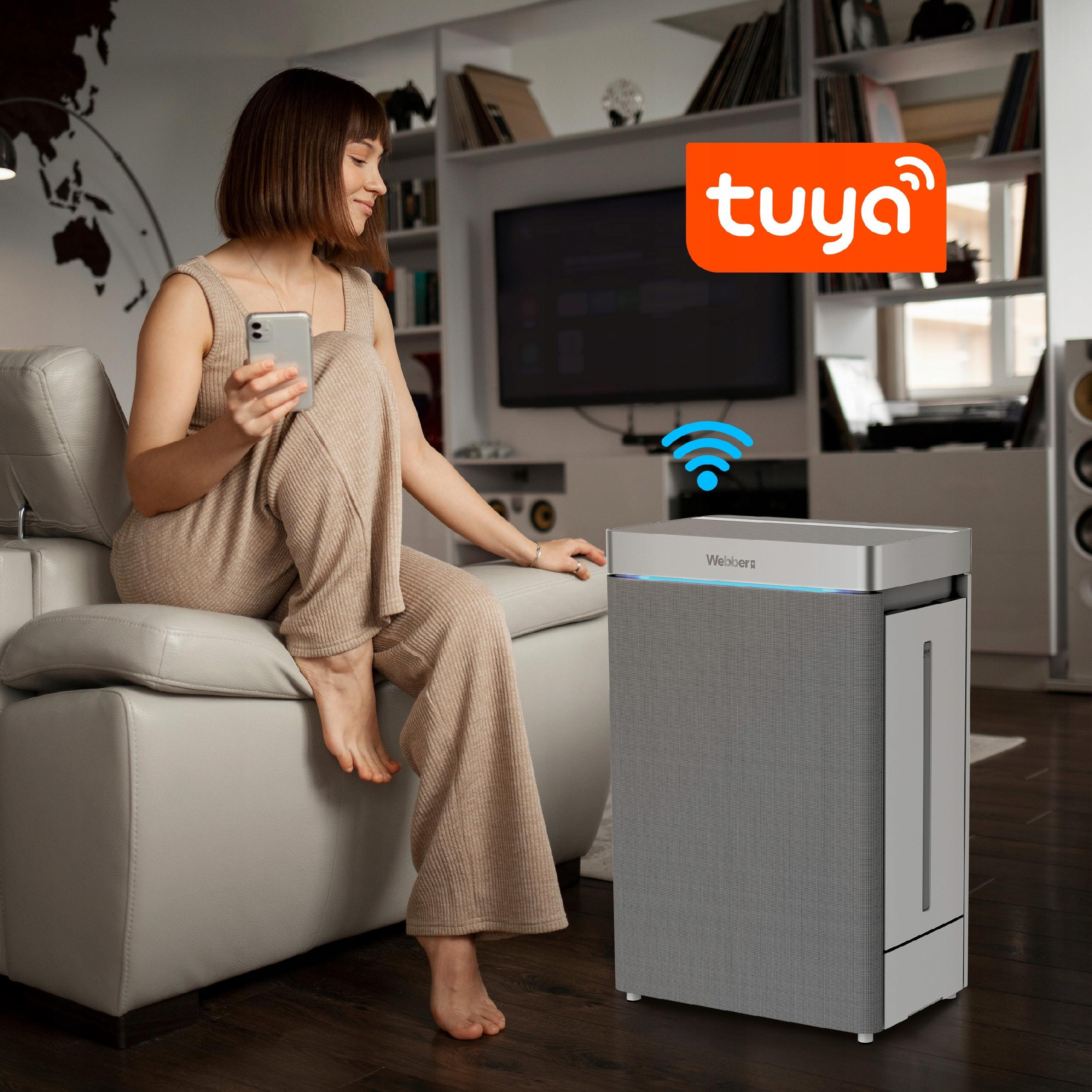 Kobieta z telefonem w pobliżu oczyszczacza powietrza. Logo Tuya w tle. Wnętrze.