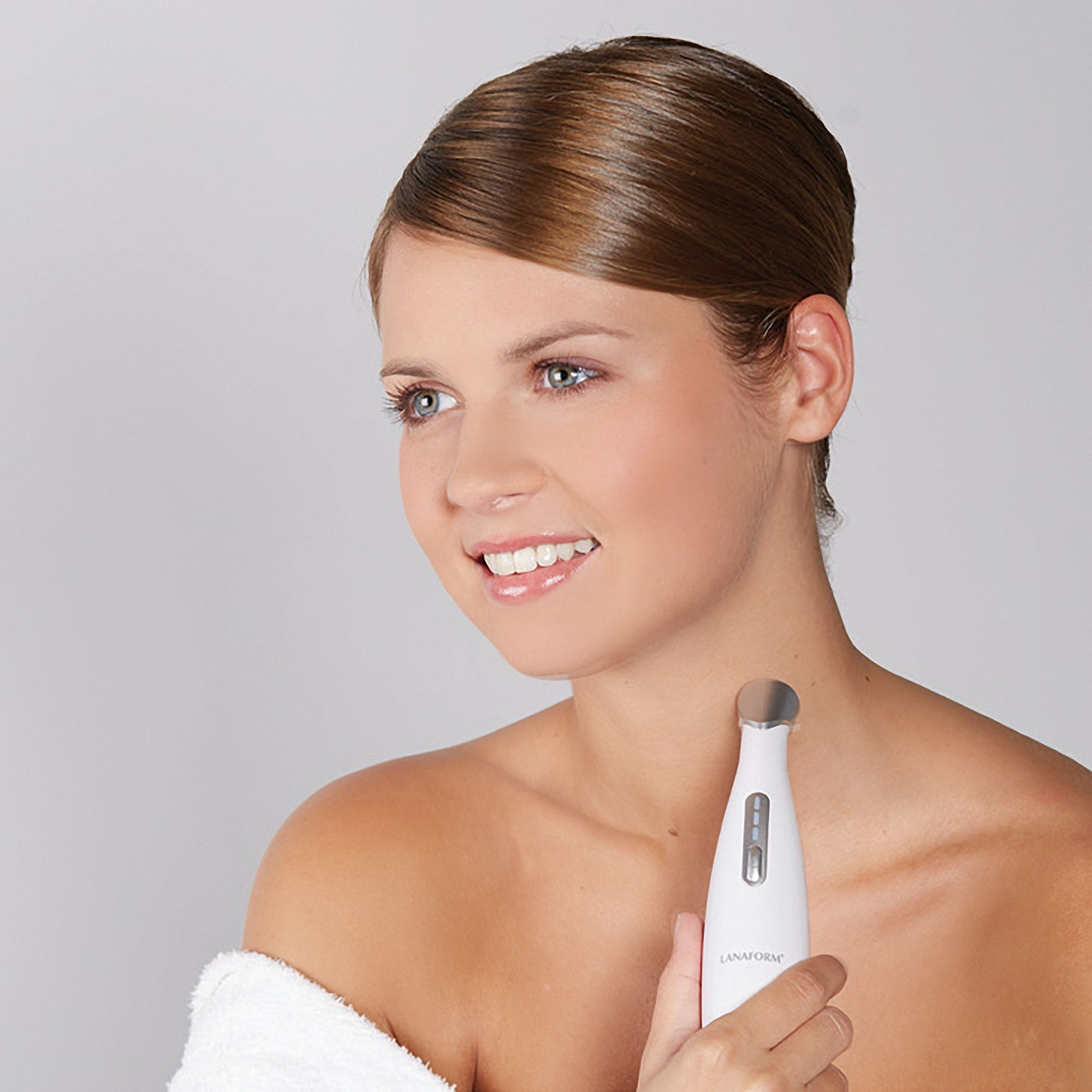 A woman smiles, holding a white device to her neck. She wears a white towel. The background is light gray.