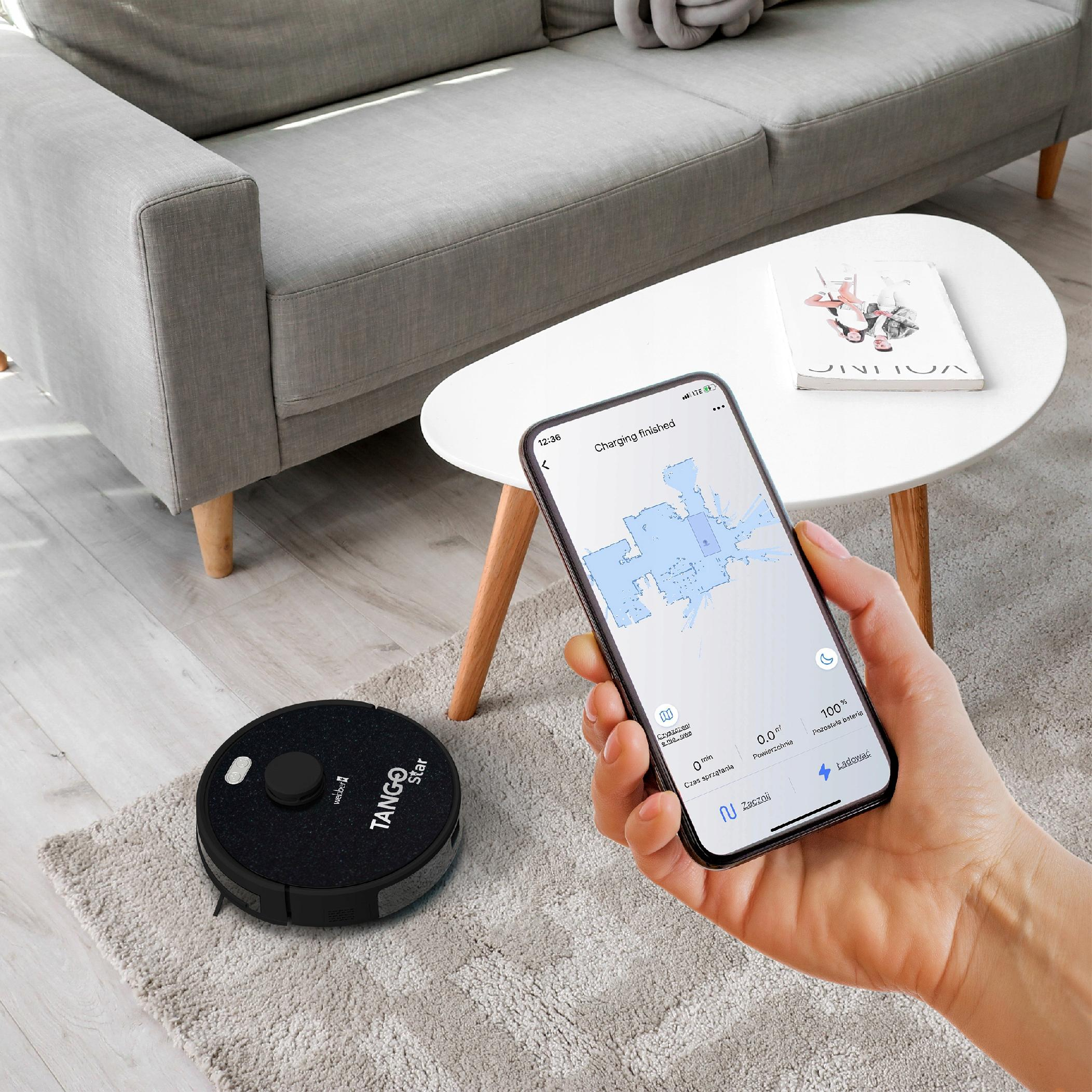 Robot sprzątający i telefon z mapą sprzątania na dywanie. W tle sofa i stolik kawowy.