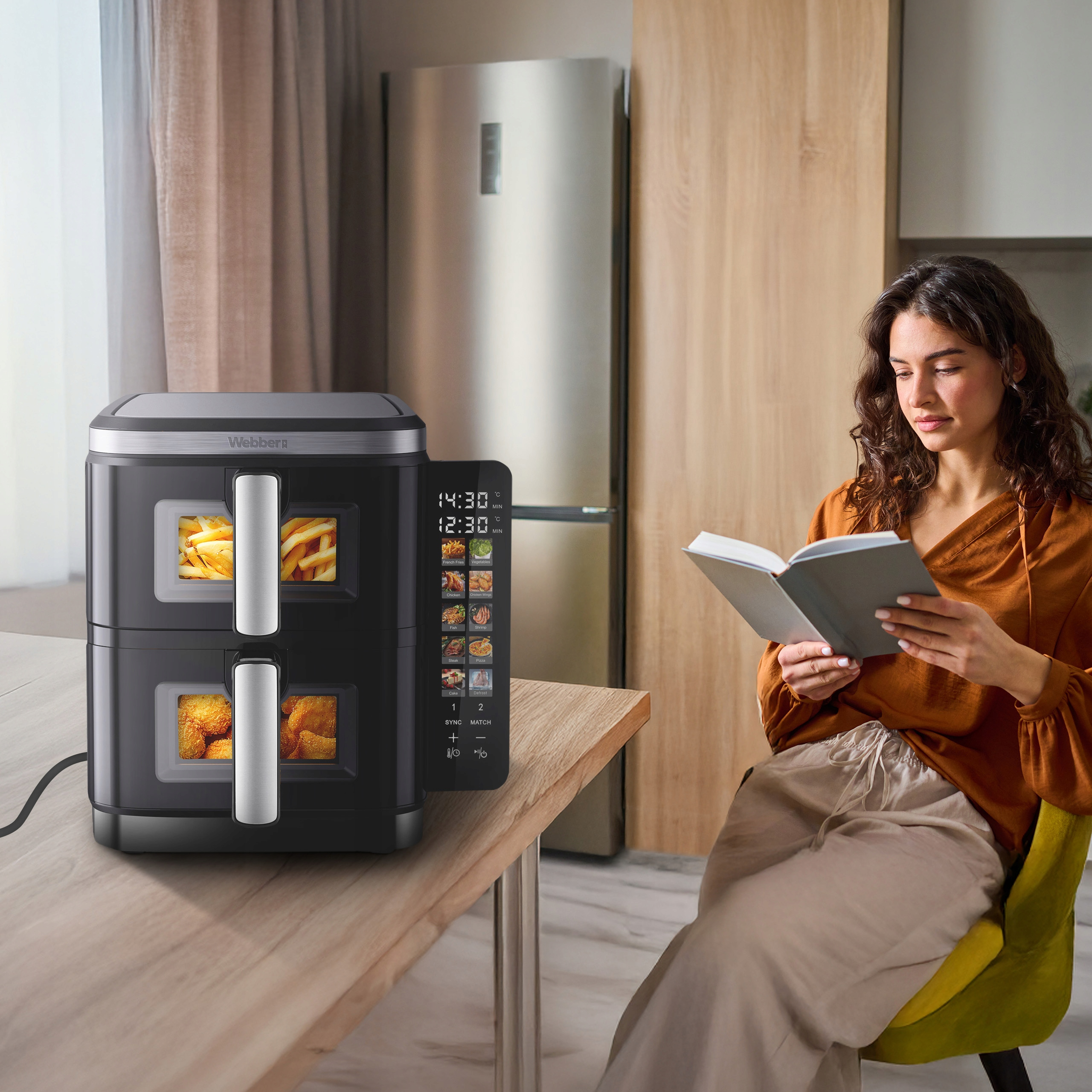 Kobieta czyta obok czarnej frytownicy powietrznej z frytkami i kurczakiem, na stole. W tle lodówka.