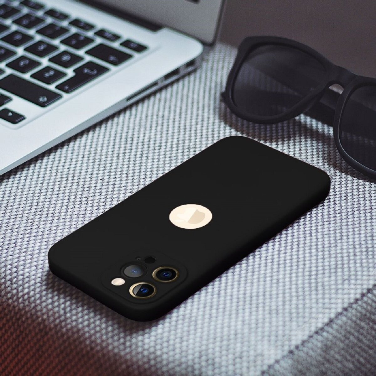Schwarzes Telefon mit goldenem Logo, Sonnenbrille und Laptop auf grauem Stoff. Das Telefon hat drei Kameralinsen.