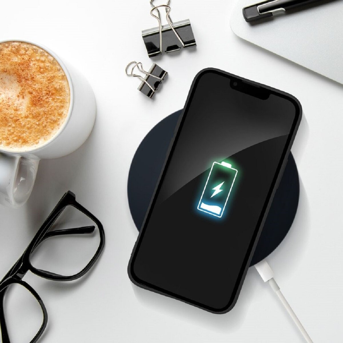 Ein Telefon auf einem kabellosen Ladegerät, das lädt. Daneben liegen eine Kaffeetasse, eine Brille und Büroklammern auf einer weißen Oberfläche.