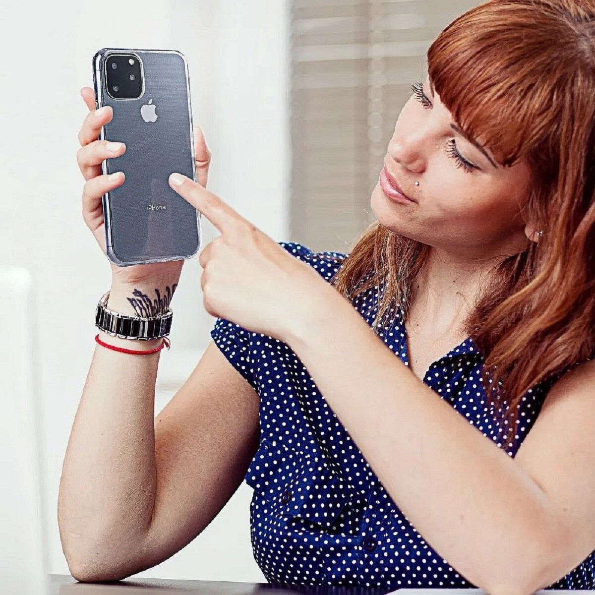 Eine Frau zeigt auf ein iPhone in ihrer Hand. Sie trägt ein gepunktetes Hemd und hat rotes Haar.