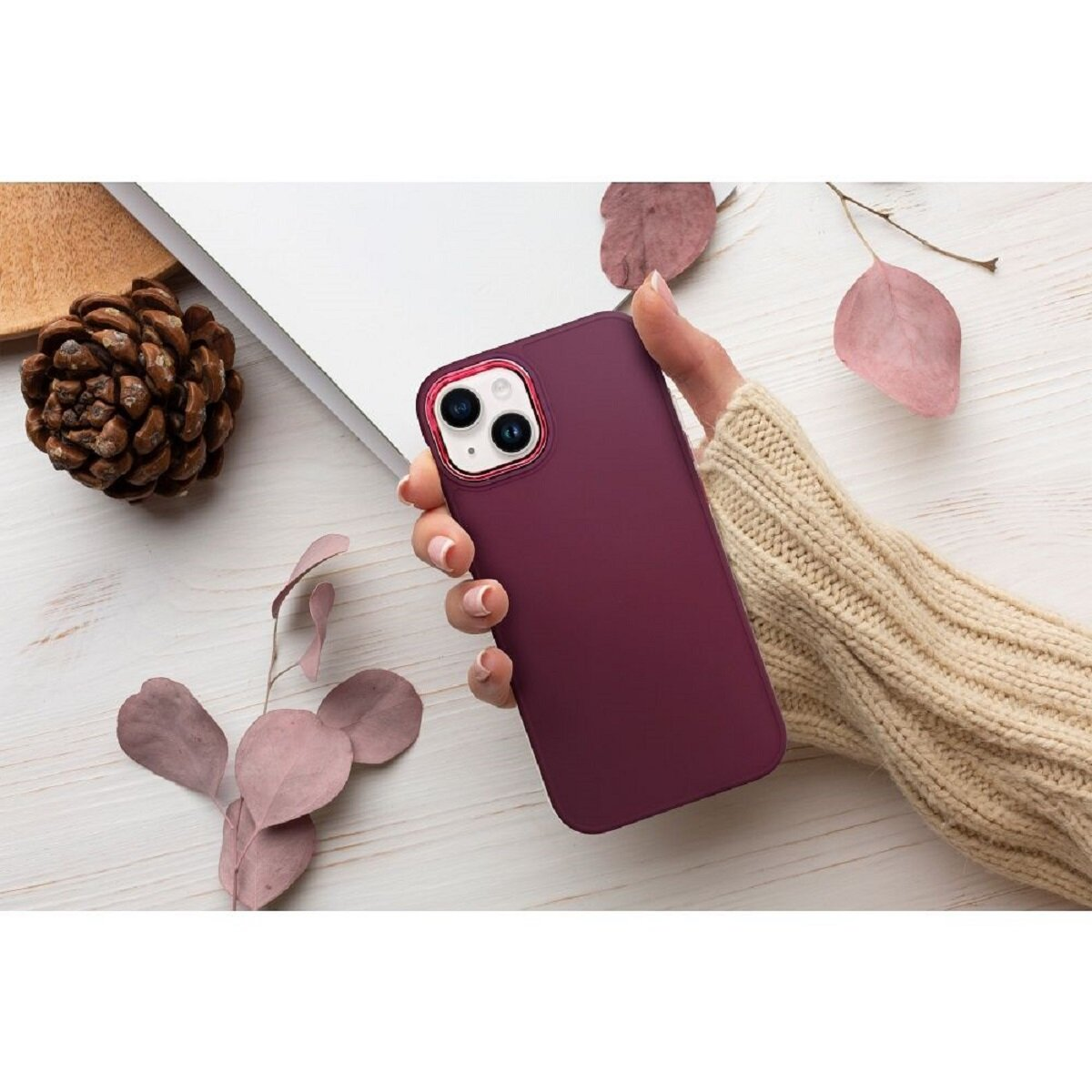 Eine Hand hält ein weinrotes Telefon mit goldenem Kameraring. Tannenzapfen und Blätter liegen daneben, mit weißem und Holz Hintergrund.