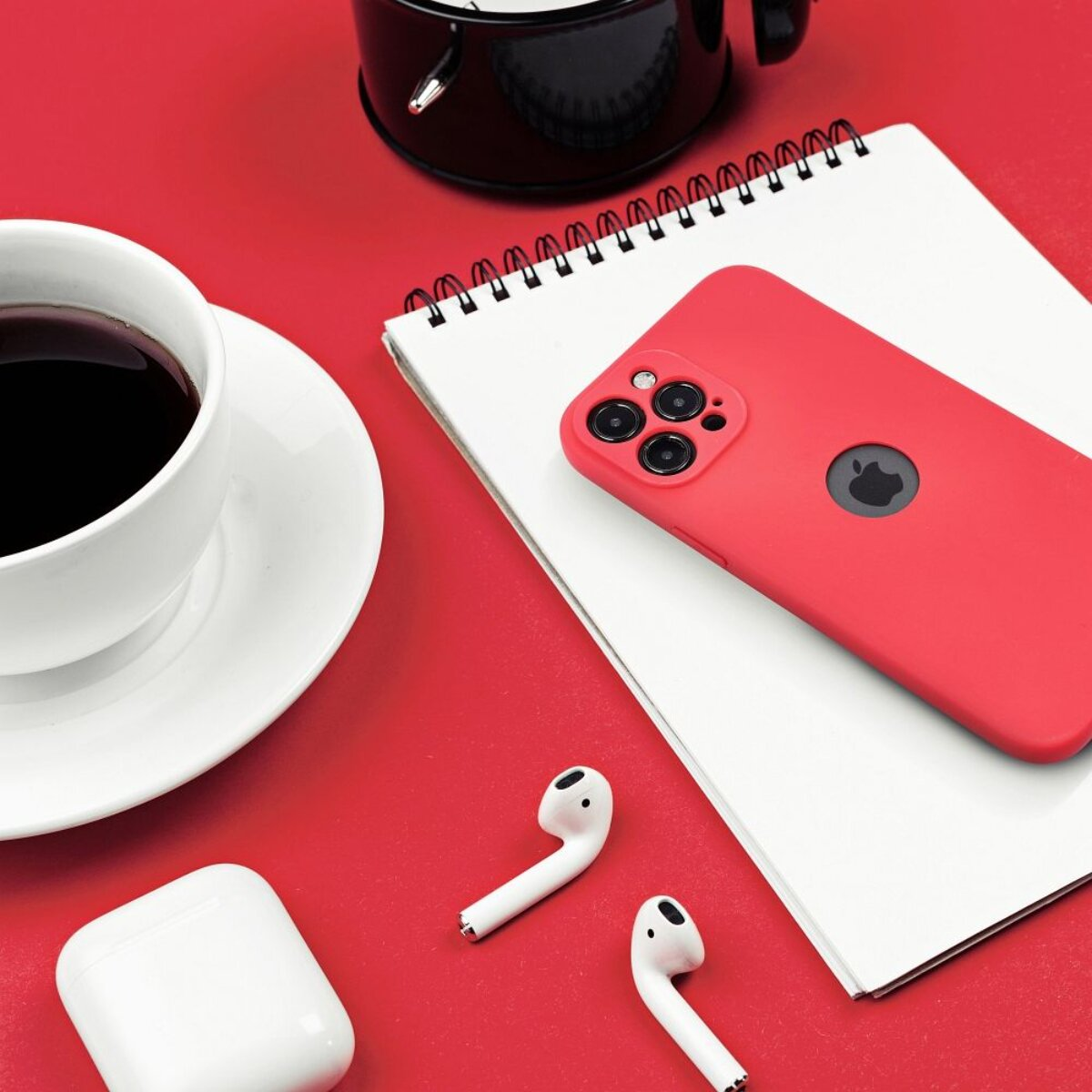 Eine rote Handyhülle, ein weißes Notizbuch, eine Kaffeetasse, Ohrhörer und ein Ladeetui auf roter Oberfläche.