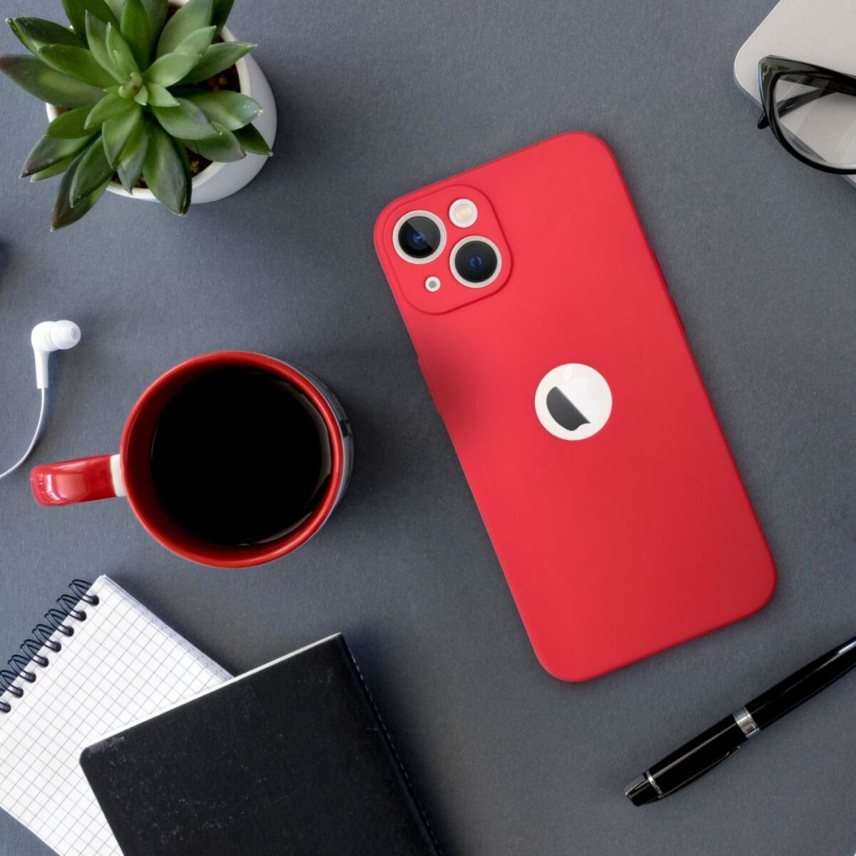Ein rotes iPhone liegt auf einem Schreibtisch neben einer Kaffeetasse, einem Notizbuch, einem Stift und einer Pflanze. Kopfhörer und Brille sind ebenfalls vorhanden.
