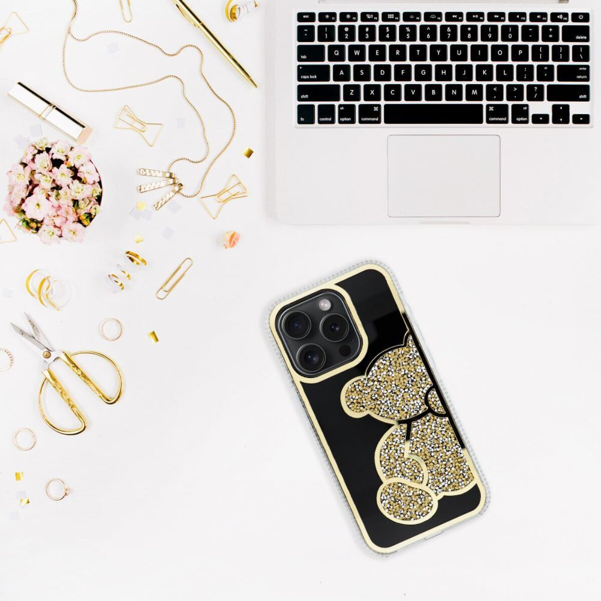 Ein Telefon mit Bärenhülle, Laptop, Zubehör und Blumen auf weißer Oberfläche. Gold- und Rosatöne dominieren.