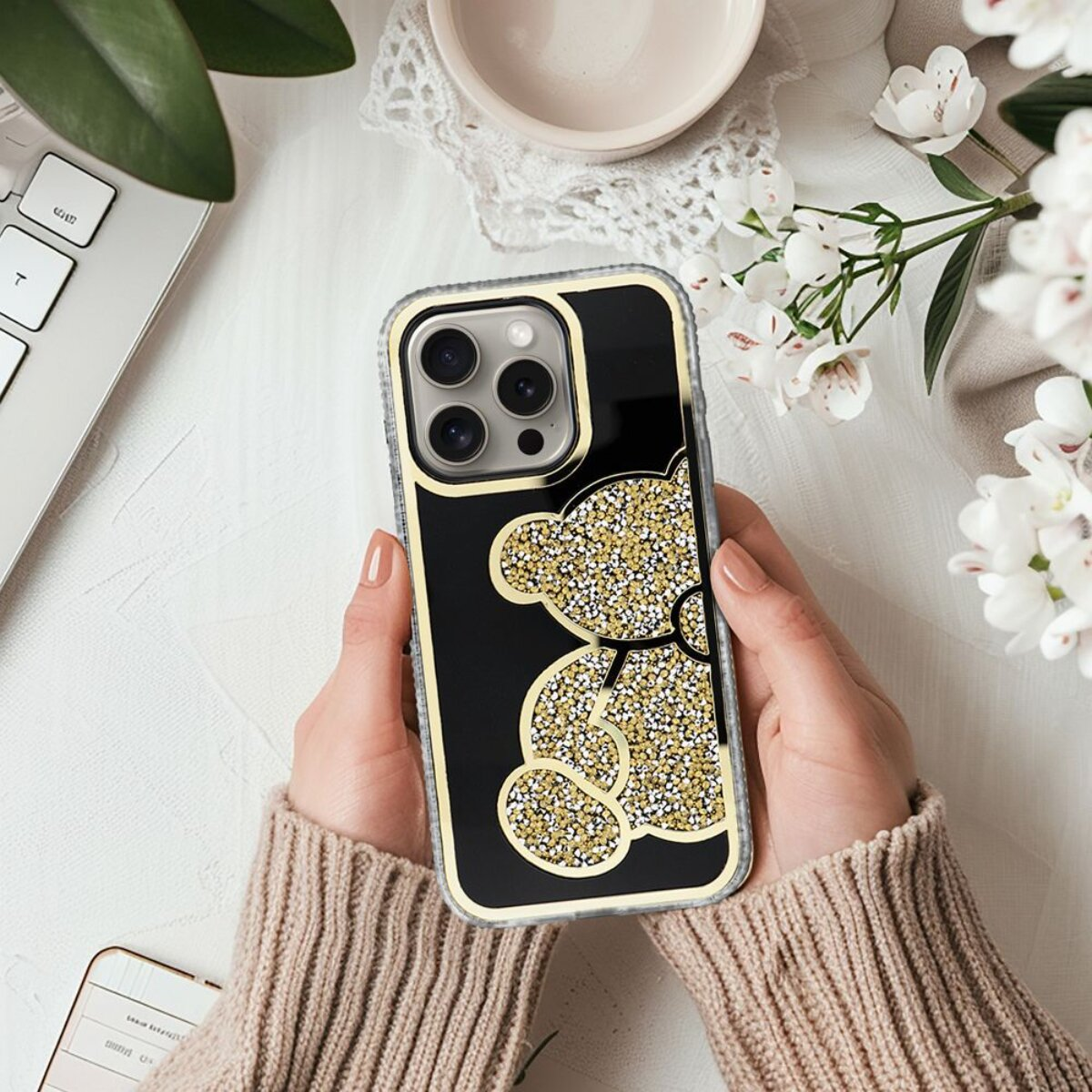 Hände halten eine Handyhülle mit Teddybären-Design, umgeben von Blumen, einer Tastatur und einem Notizbuch auf weißer Oberfläche.