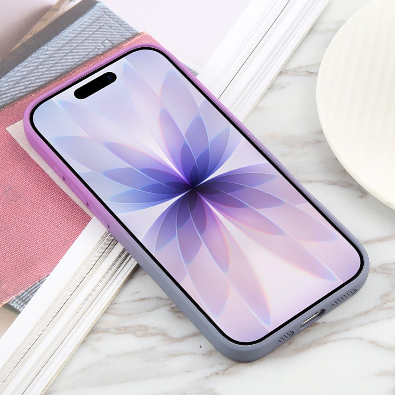Ein Smartphone mit lila-grauer Hülle, das ein Blumengemälde zeigt. Es liegt auf Marmor mit Büchern und einem Teller.