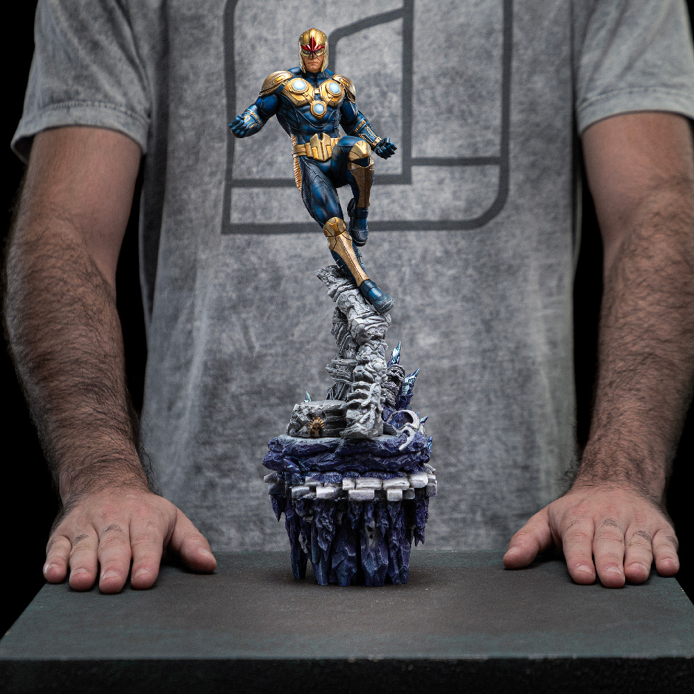 Un uomo tiene in mano una statua di Nova. La statua raffigura un supereroe in tuta blu e oro, in posa su una base rocciosa.