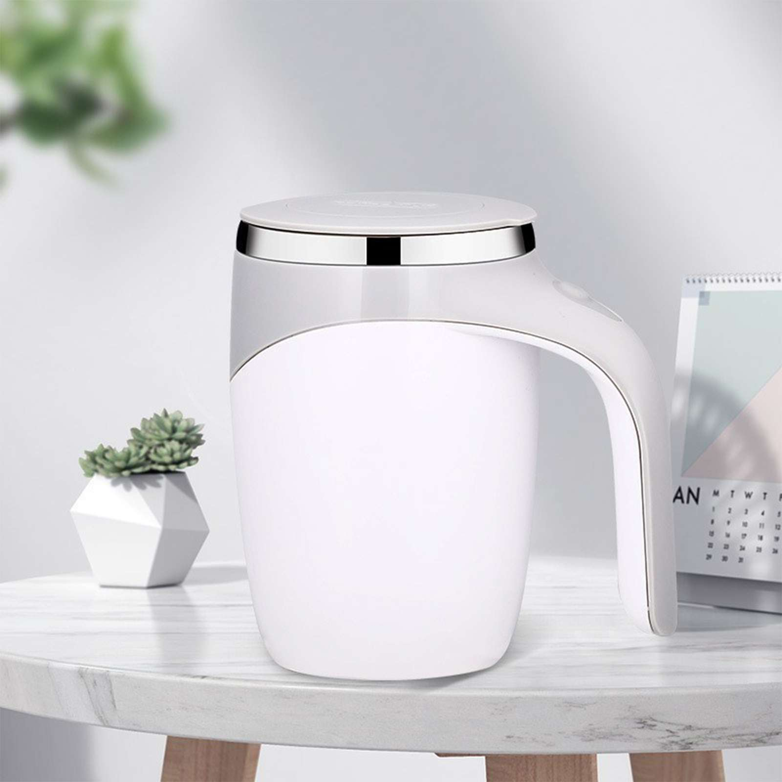 Ein weißer selbstquirlender Becher mit Griff steht auf einem Tisch. Eine kleine Sukkulentenpflanze und ein Kalender sind ebenfalls vorhanden.