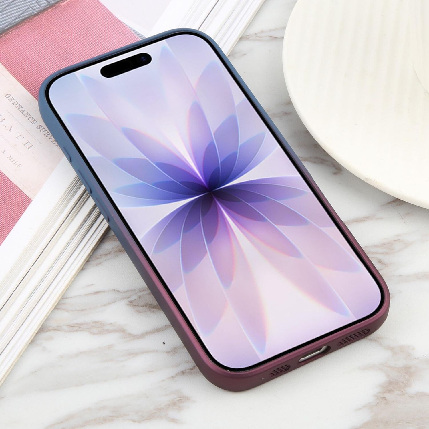 Ein Telefon in einer Hülle zeigt ein Blumenmuster. Es liegt auf einer Marmoroberfläche, neben einem Buch und einem weißen Teller.