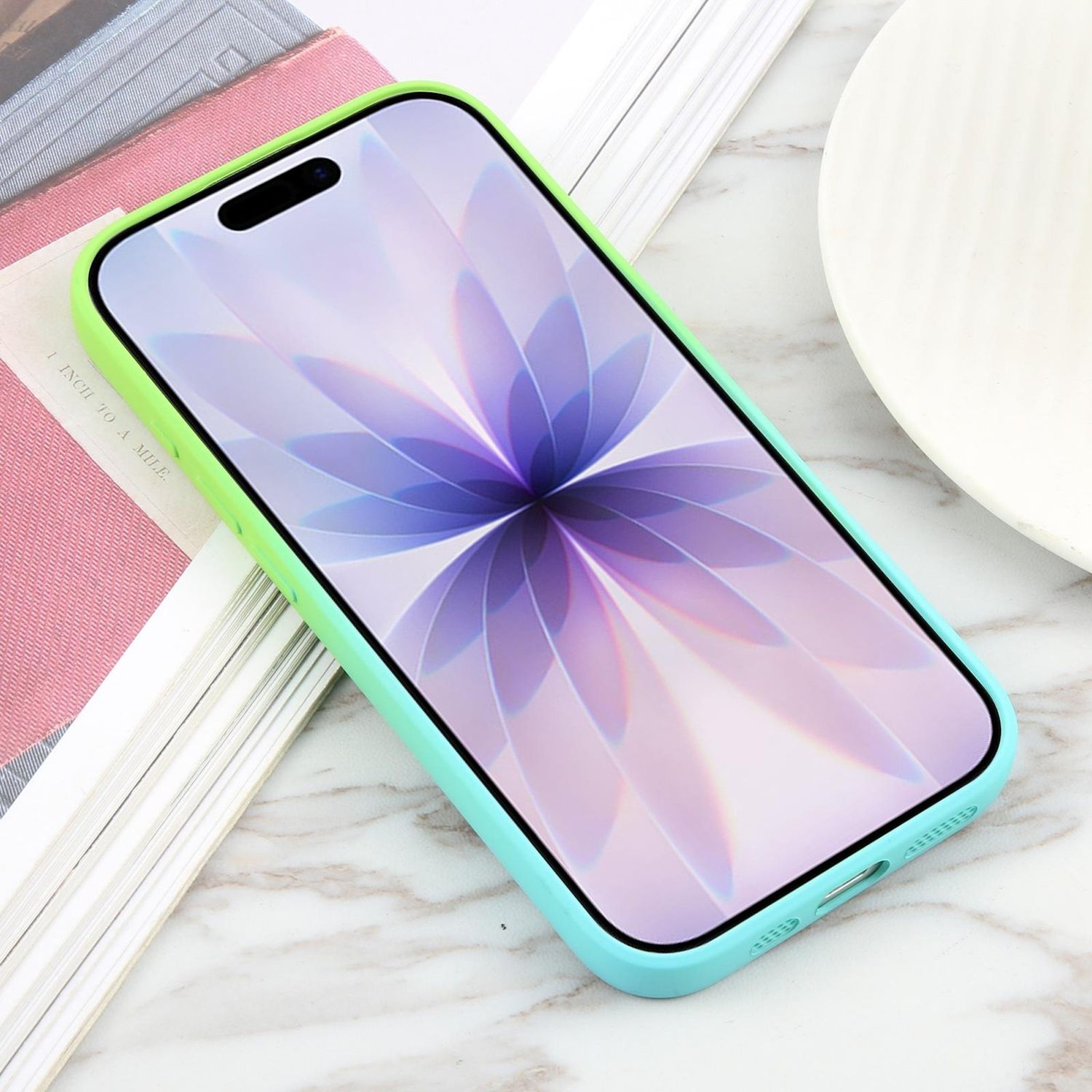 Ein Telefon in einer Hülle mit Farbverlauf von Grün nach Blau. Der Bildschirm zeigt eine Blume. Das Telefon liegt auf Büchern und einer Marmoroberfläche.