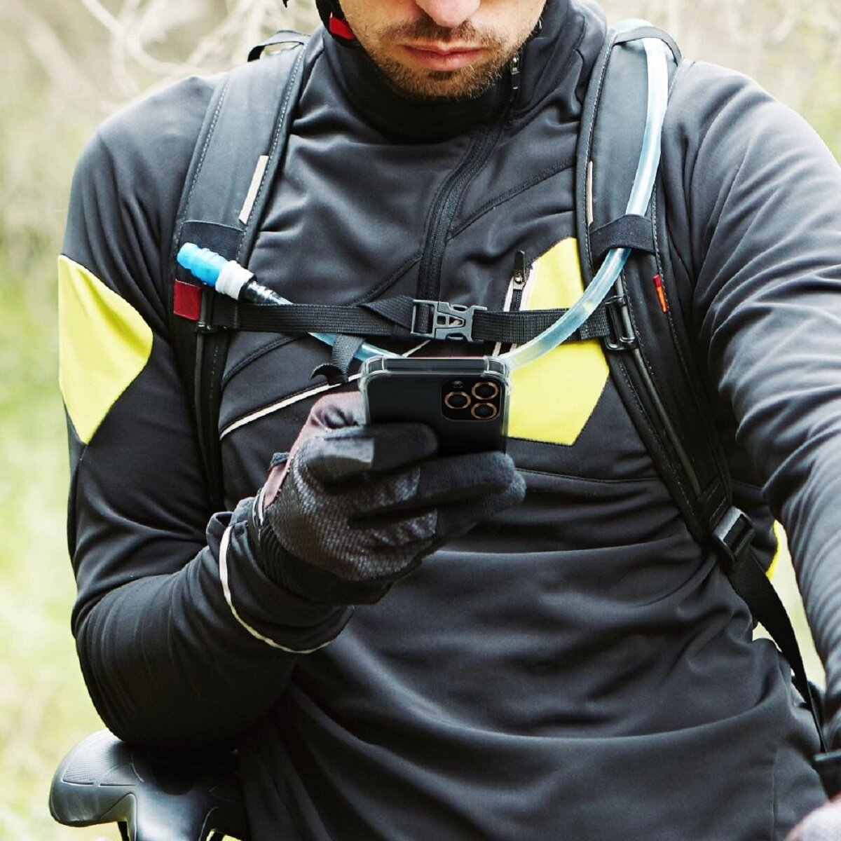 Radfahrer in schwarzer Jacke benutzt Telefon, trägt Handschuhe und Rucksack mit Trinksystem. Gelbe Akzente auf der Jacke.
