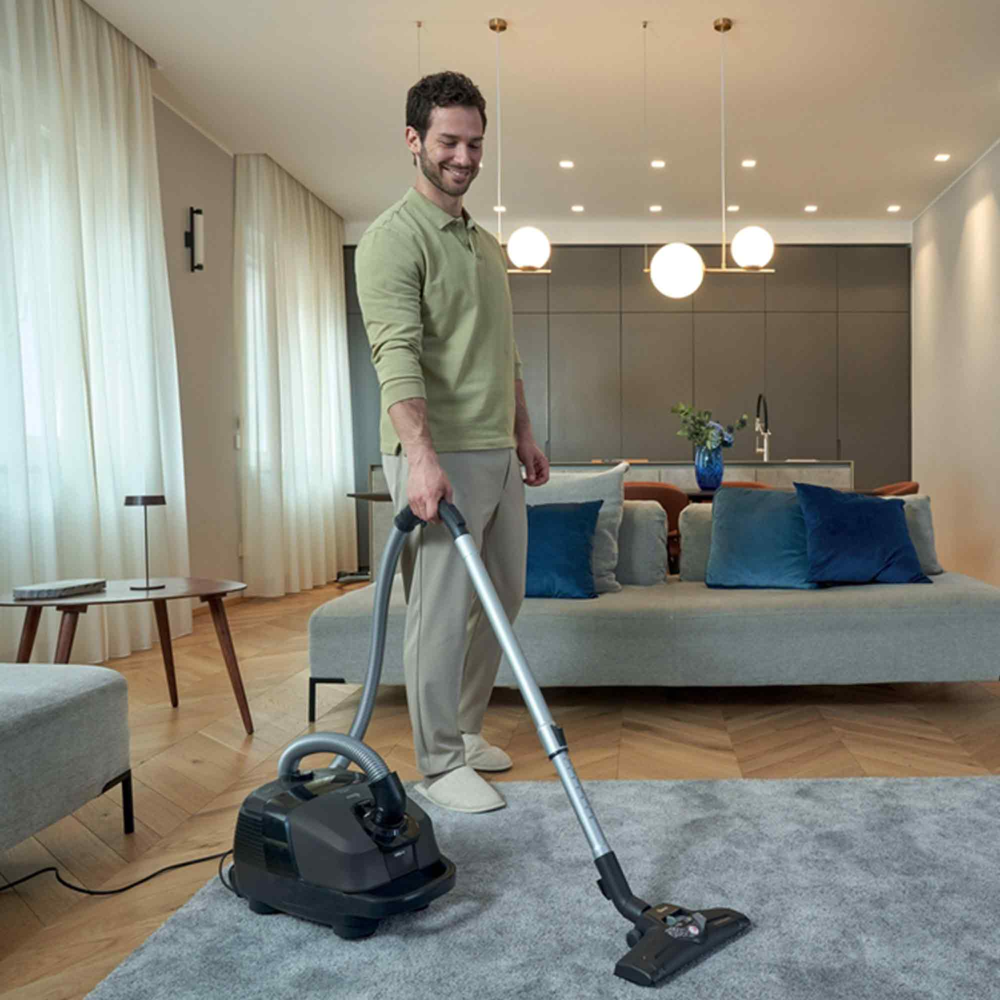 Mann, der einen Teppich im Wohnzimmer saugt. Im Hintergrund sind ein Sofa, Vorhänge und Lampen.