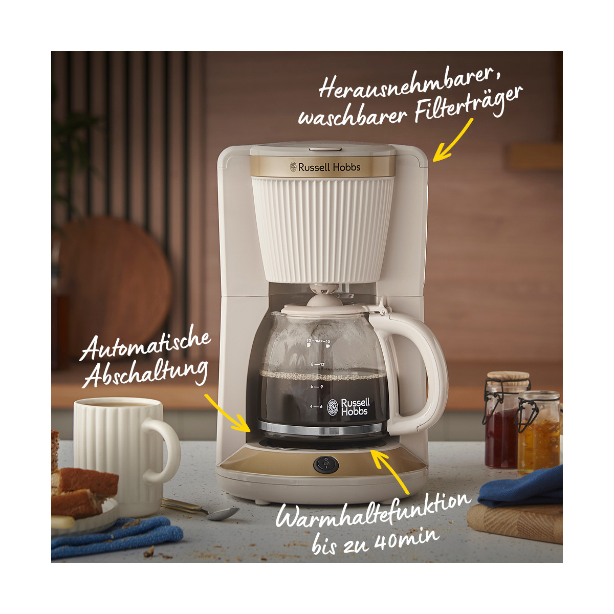 Una macchina da caffè Russell Hobbs bianca e oro con una caraffa di vetro, una tazza e alcuni barattoli su un bancone.