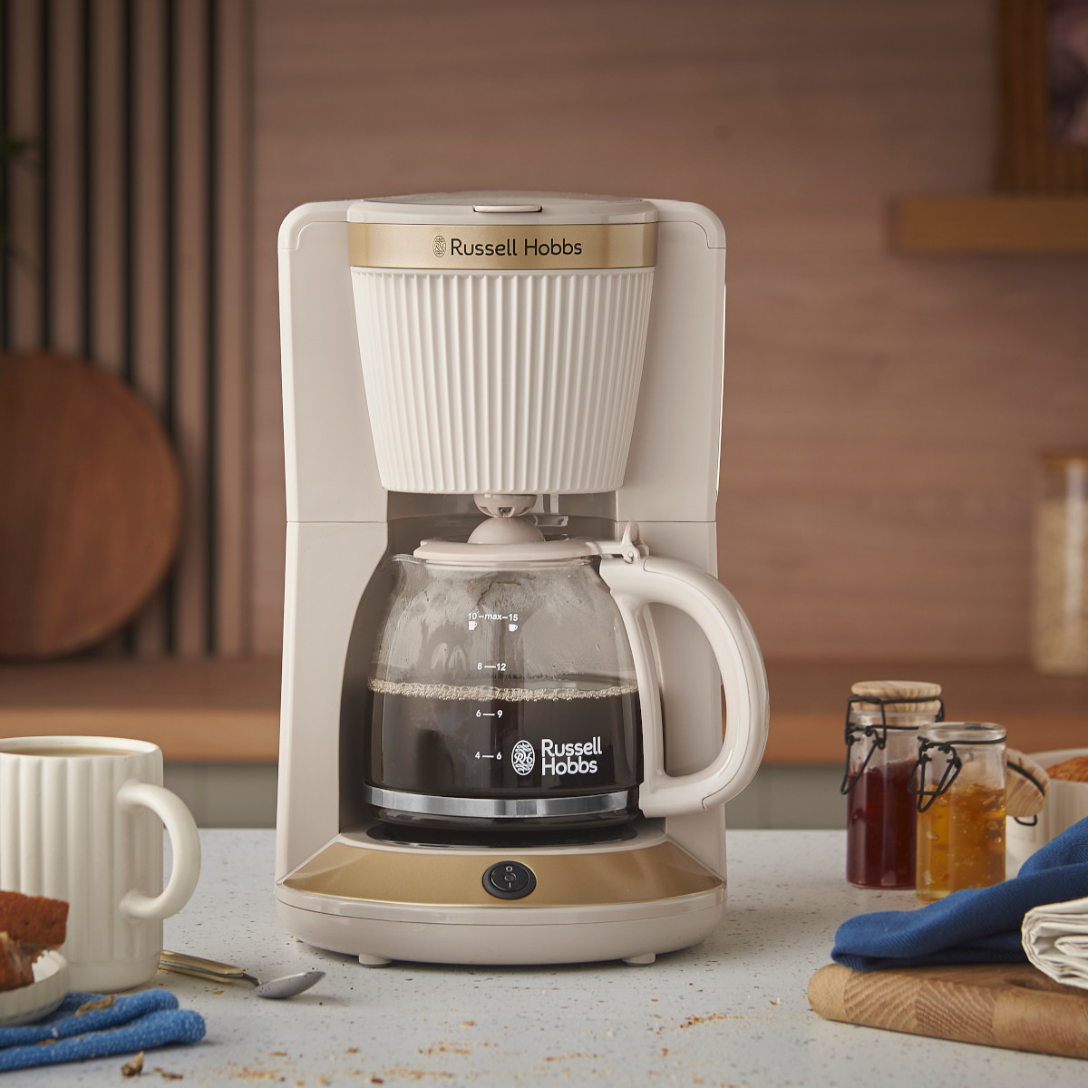 Una caffettiera Russell Hobbs con una caraffa in vetro piena di caffè. Una tazza, pane e barattoli di marmellata in primo piano.