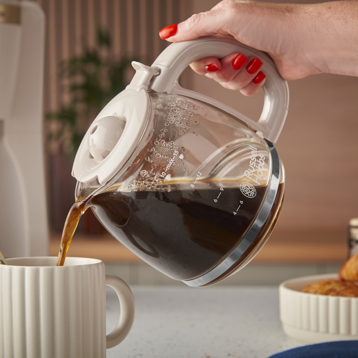 Eine Hand gießt Kaffee aus einer Glaskaraffe in eine weiße Tasse. Im Hintergrund befindet sich eine Kaffeemaschine. Rote Nägel sind sichtbar.