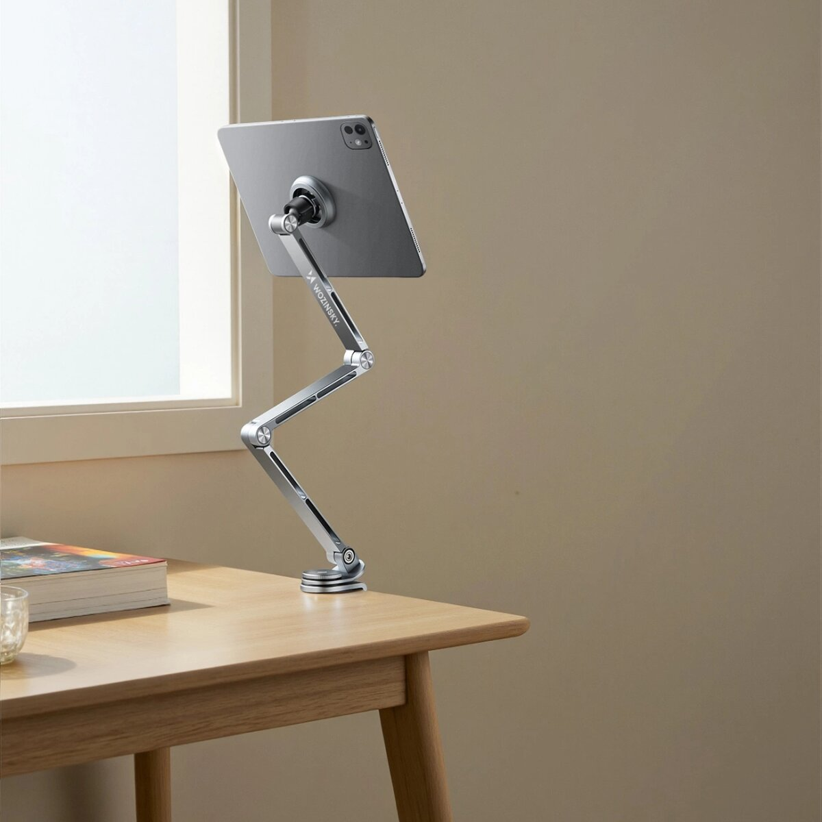 Een tablet is gemonteerd op een zilveren standaard op een houten tafel. Een raam en een boek staan ook op de tafel. De achtergrond is beige.