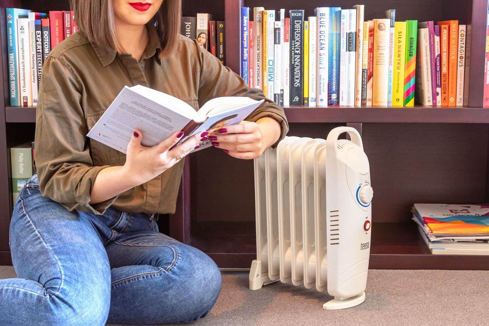 Kobieta z czerwoną szminką czyta książkę siedząc obok białego grzejnika. Regały z książkami w tle.