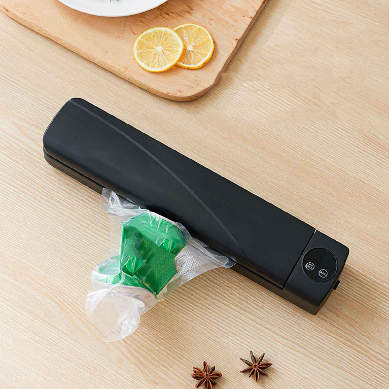 Ein schwarzes Vakuumiergerät mit einem grünen Gegenstand in einer Tüte liegt auf einem Holztisch. Zitronen und Gewürze sind ebenfalls auf dem Tisch.