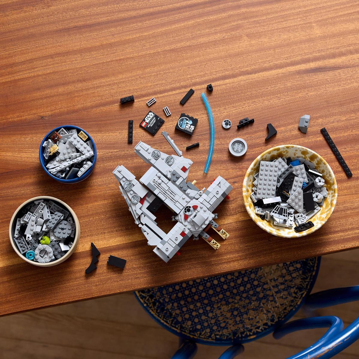 Una nave Lego Star Wars parcialmente ensamblada con piezas dispersas y cuencos de bloques sobre una mesa de madera. Una silla azul está en el fondo.